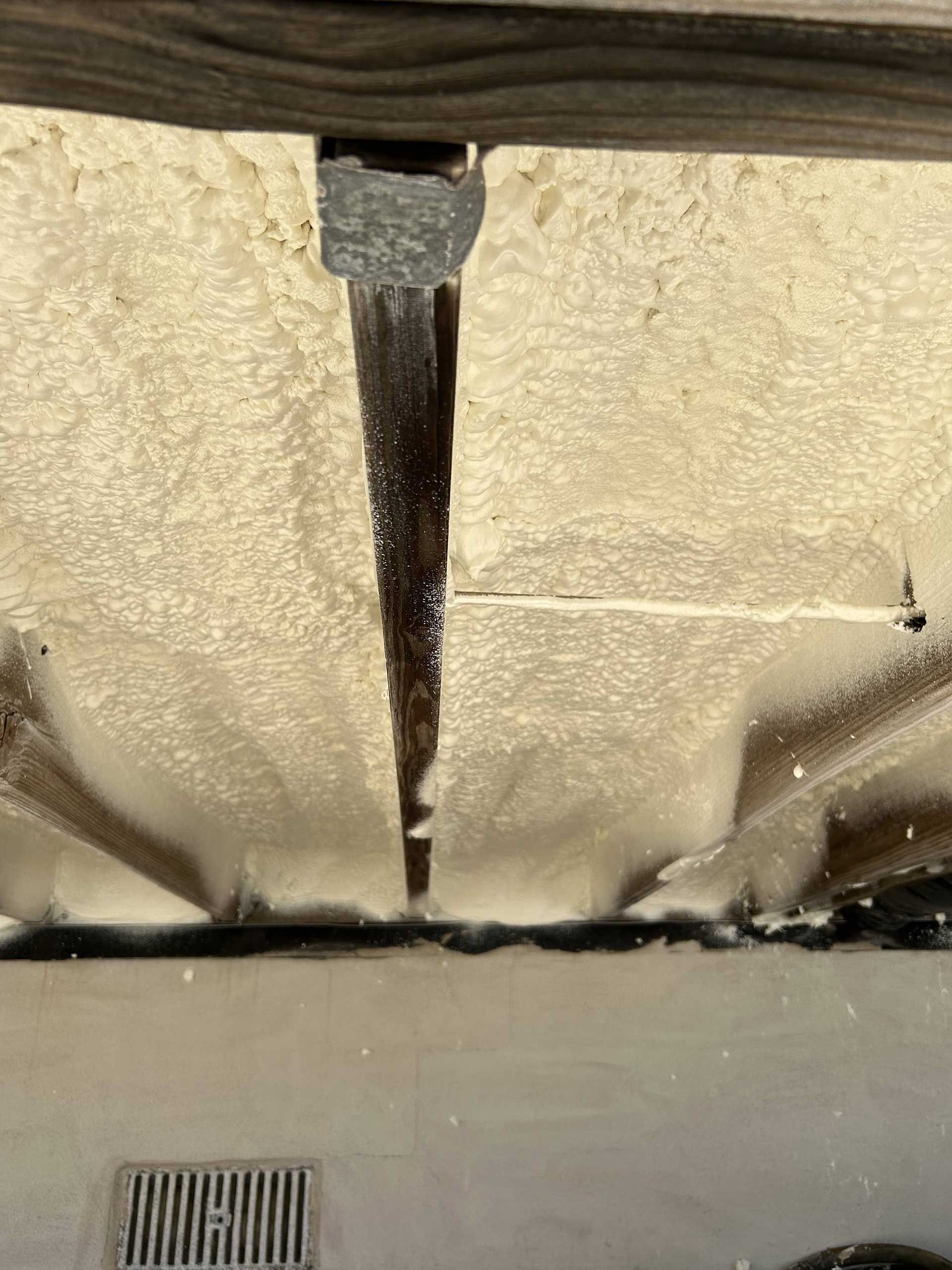 Spray foam insulation fills the gaps between wooden ceiling beams above a concrete floor with a metal drain grate.