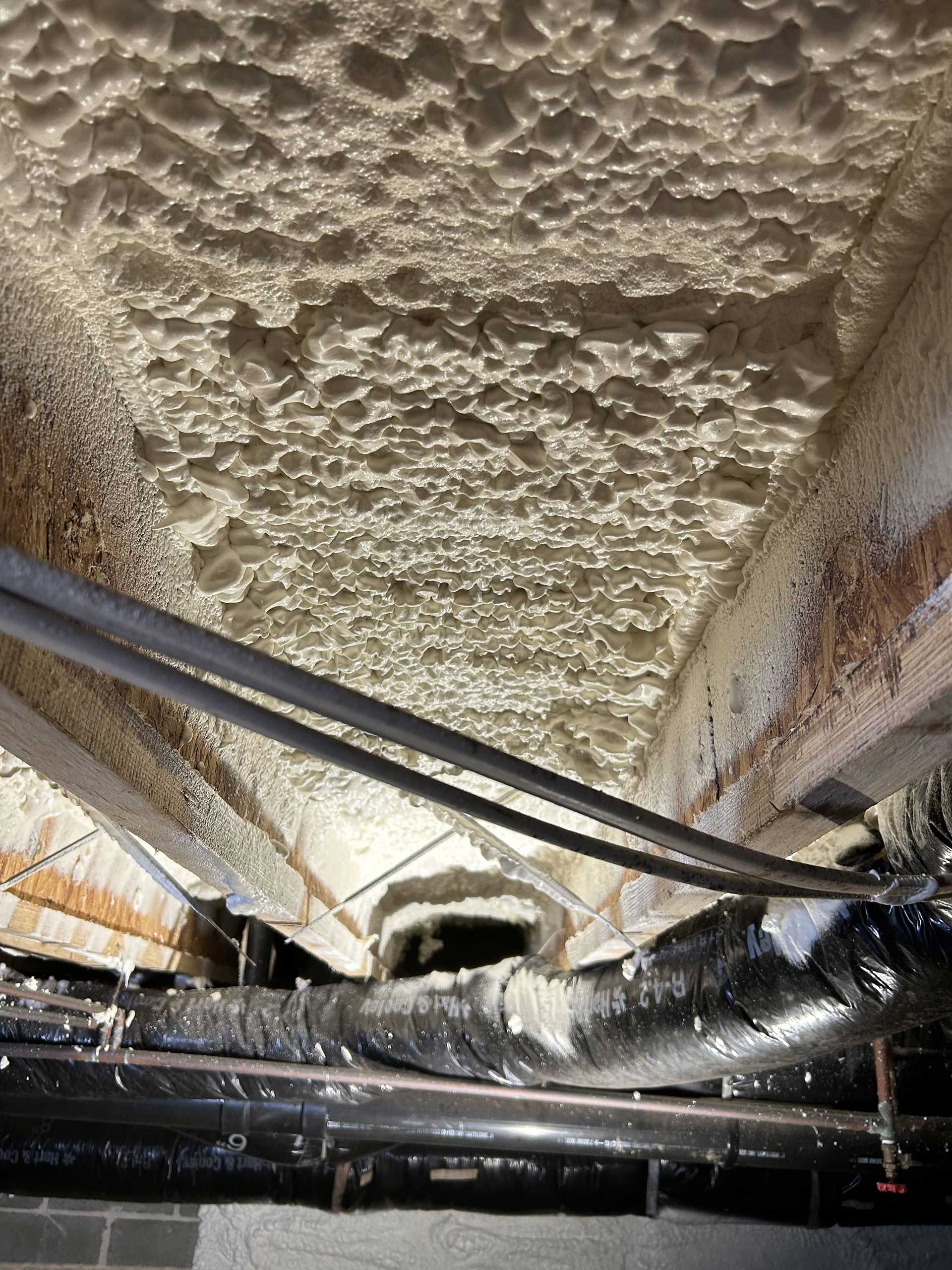 Spray foam insulation applied to the underside of floor joists in a crawl space, with pipes and ductwork visible below.