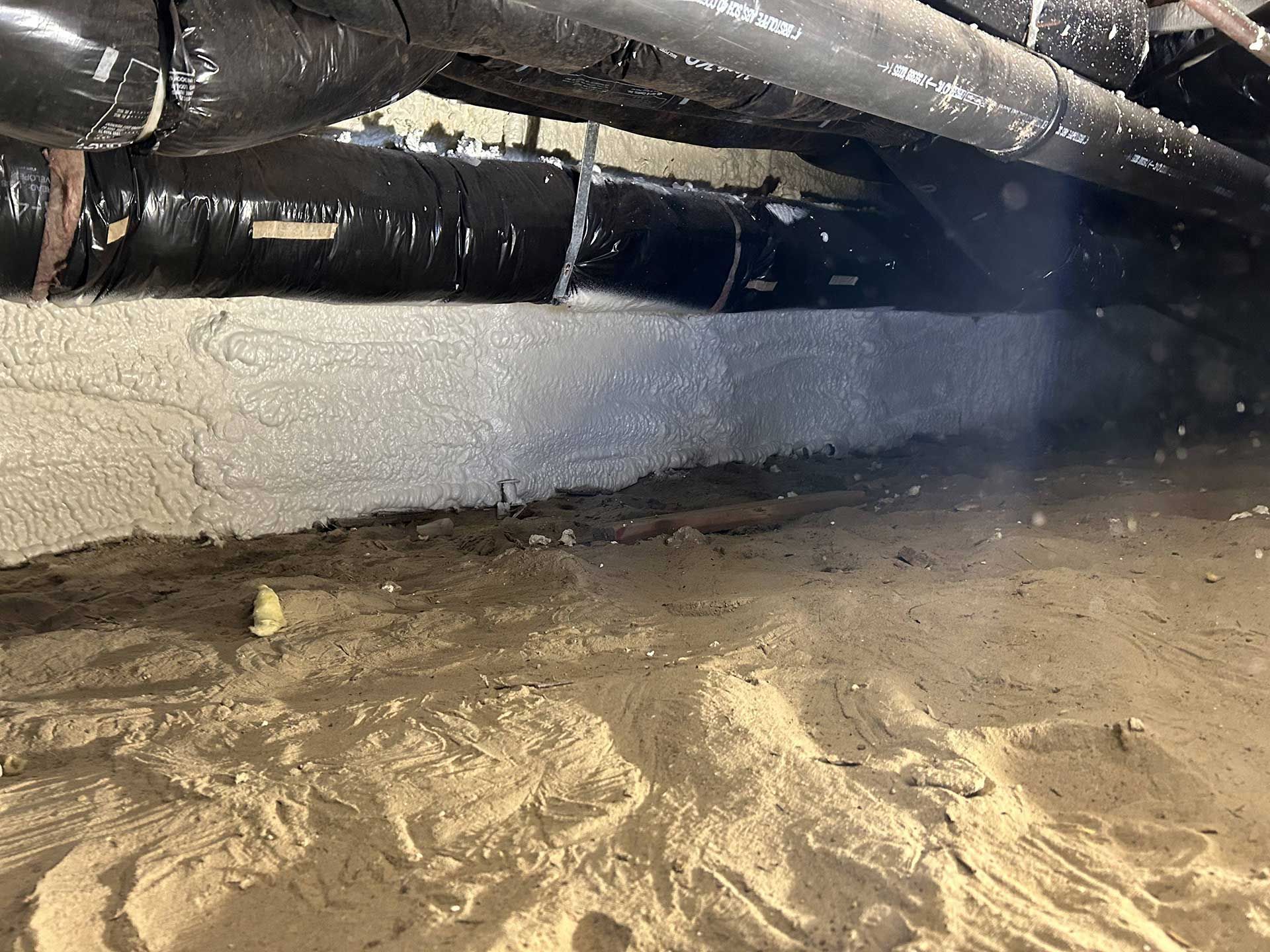 Crawl space view showing spray foam insulation applied to a concrete foundation wall beneath dark drainage pipes.