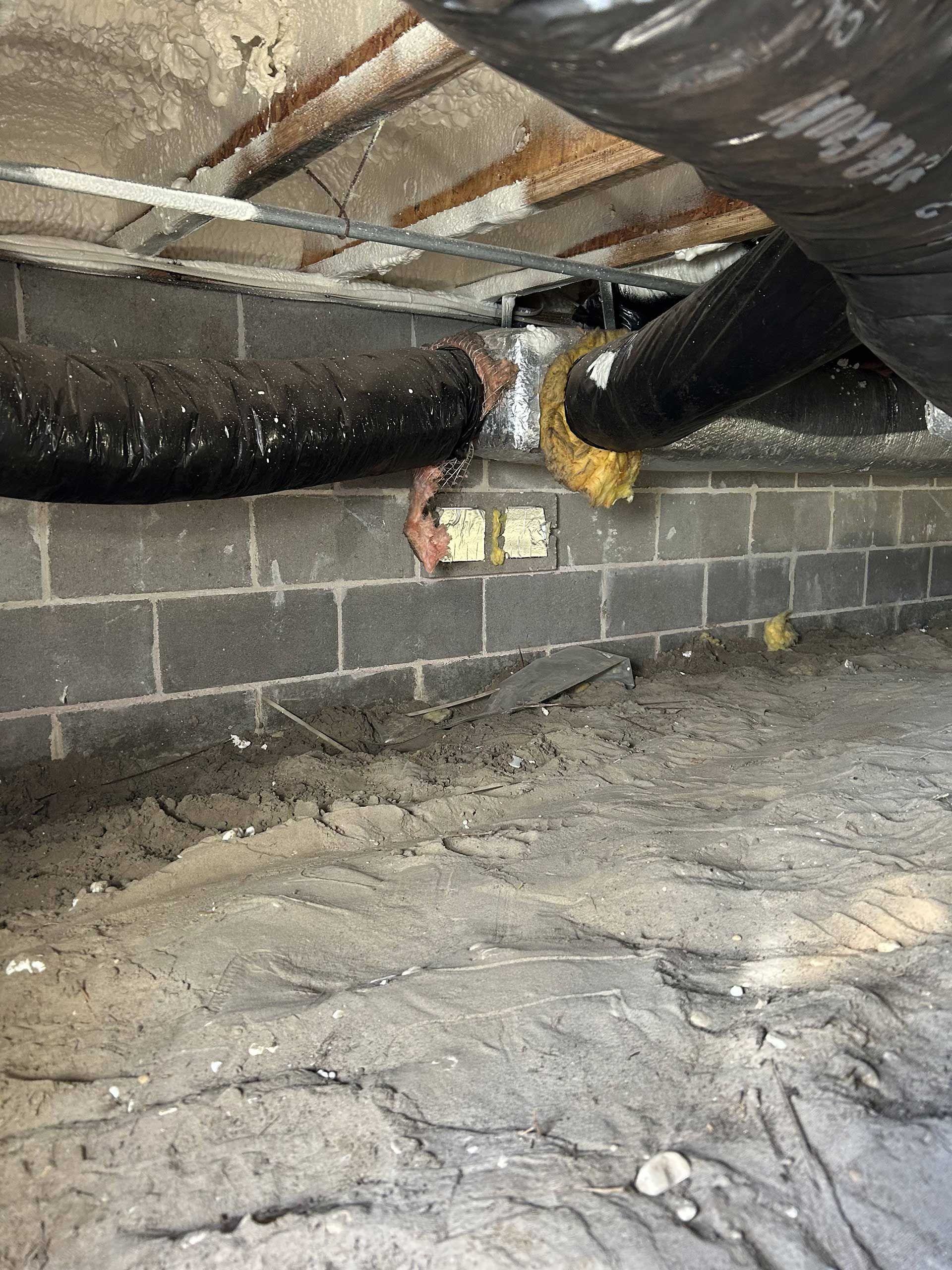 A dark crawlspace showing concrete block walls, exposed insulation, hanging black HVAC ductwork, and dirt flooring.