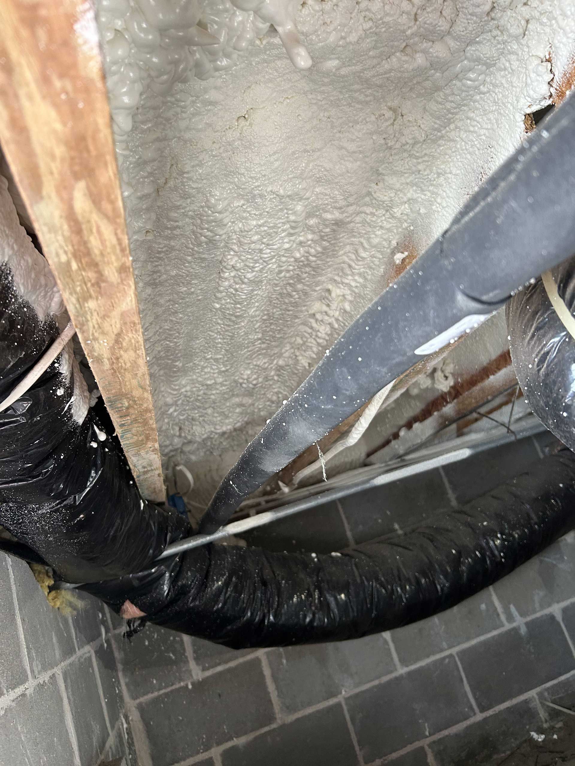 Insulated black HVAC ductwork runs along a concrete block wall beneath a floor joist with spray foam insulation.