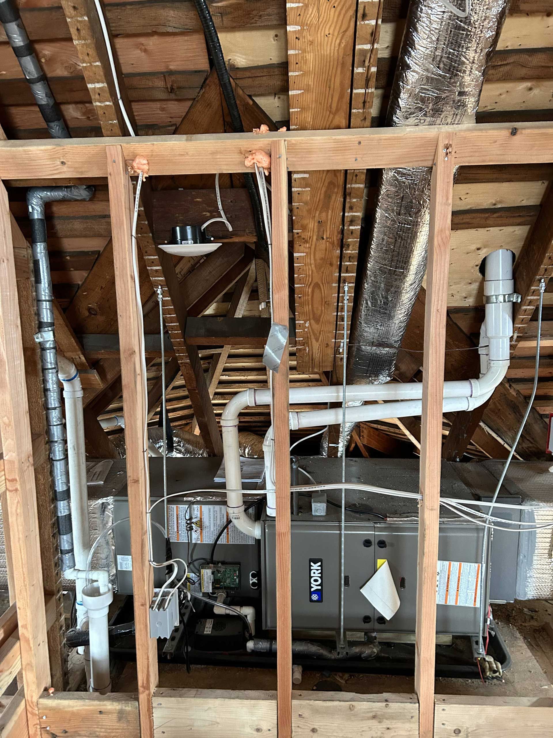 A York HVAC unit inside an unfinished wooden attic frame, with white PVC piping and silver metal ductwork visible.