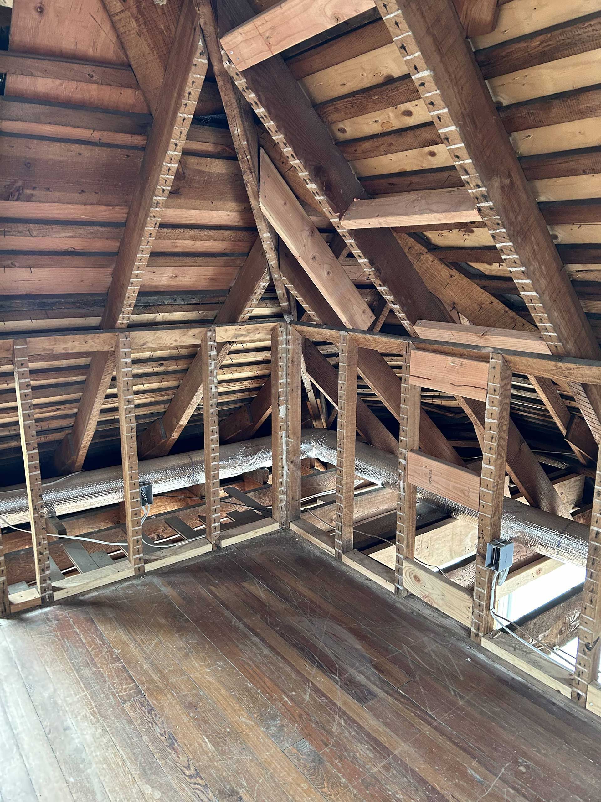 An unfinished attic space showing exposed wood roof rafters, framing for a partition wall, and dark wooden flooring.