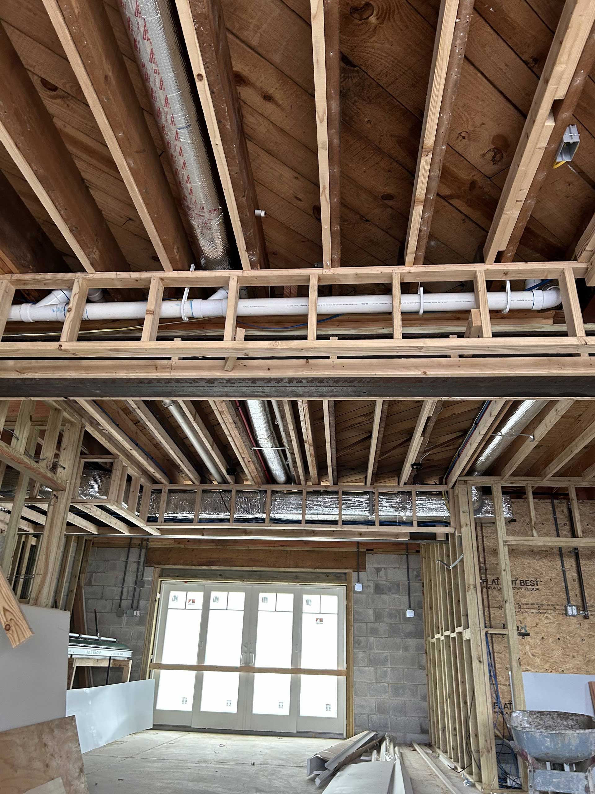 An unfinished construction site with exposed wooden ceiling joists, a white plumbing pipe, and a steel support beam.