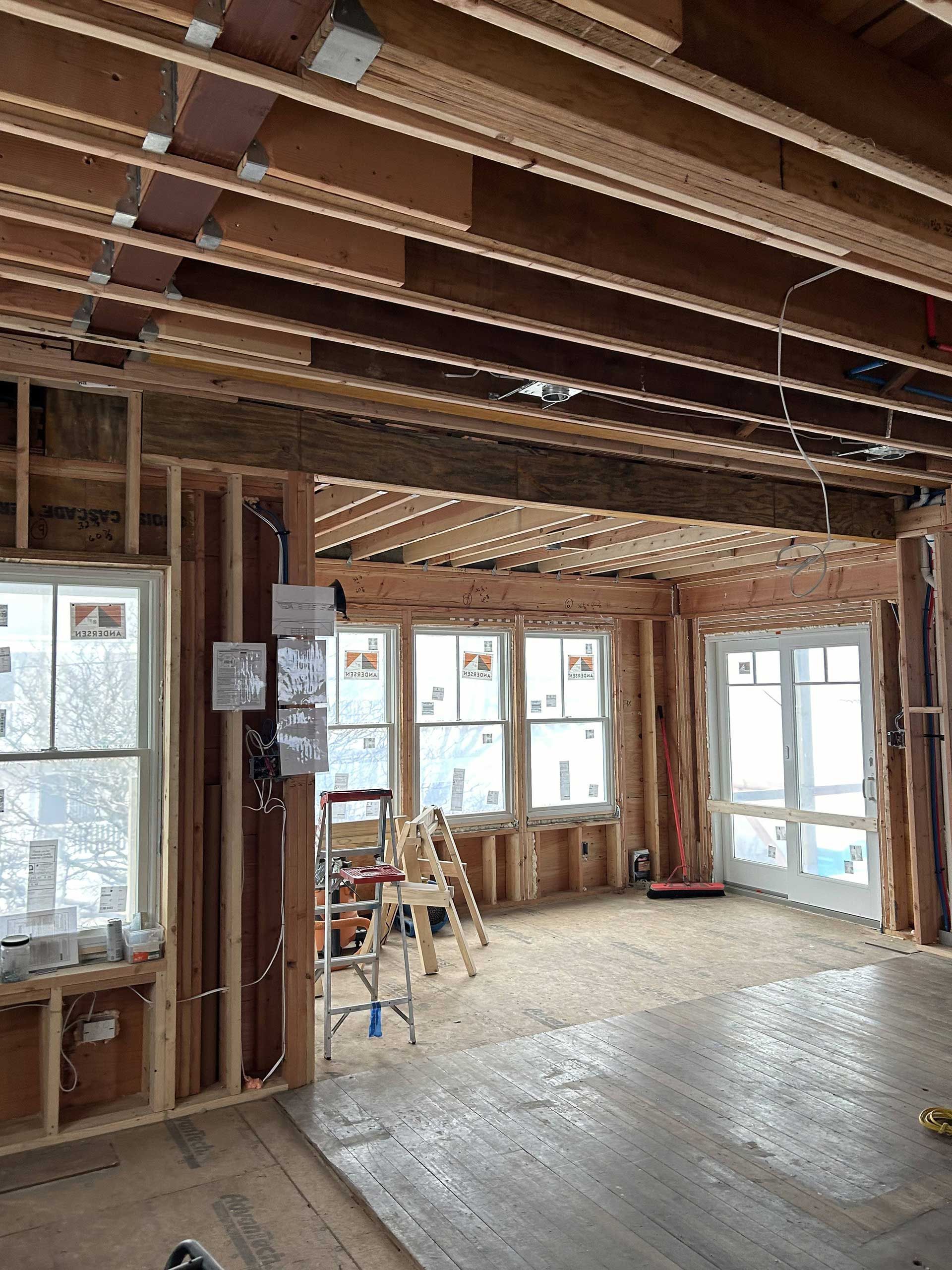 An interior view of a room under construction, featuring exposed wooden framing, subflooring, and new windows and doors.