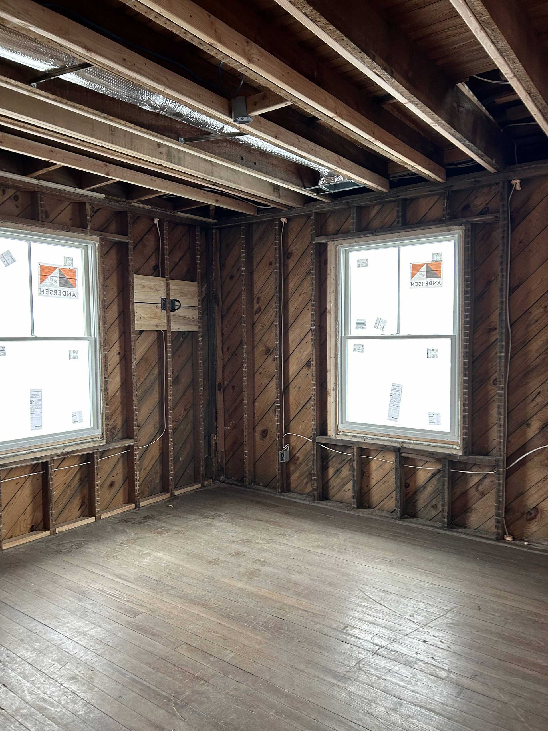 An unfinished room with exposed wooden wall studs and ceiling joists, featuring two new windows with installation labels.