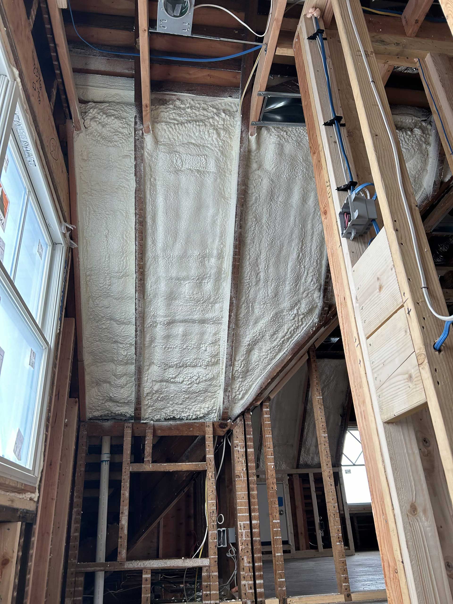 An interior view of a room under construction, showing spray foam insulation between wooden wall studs.