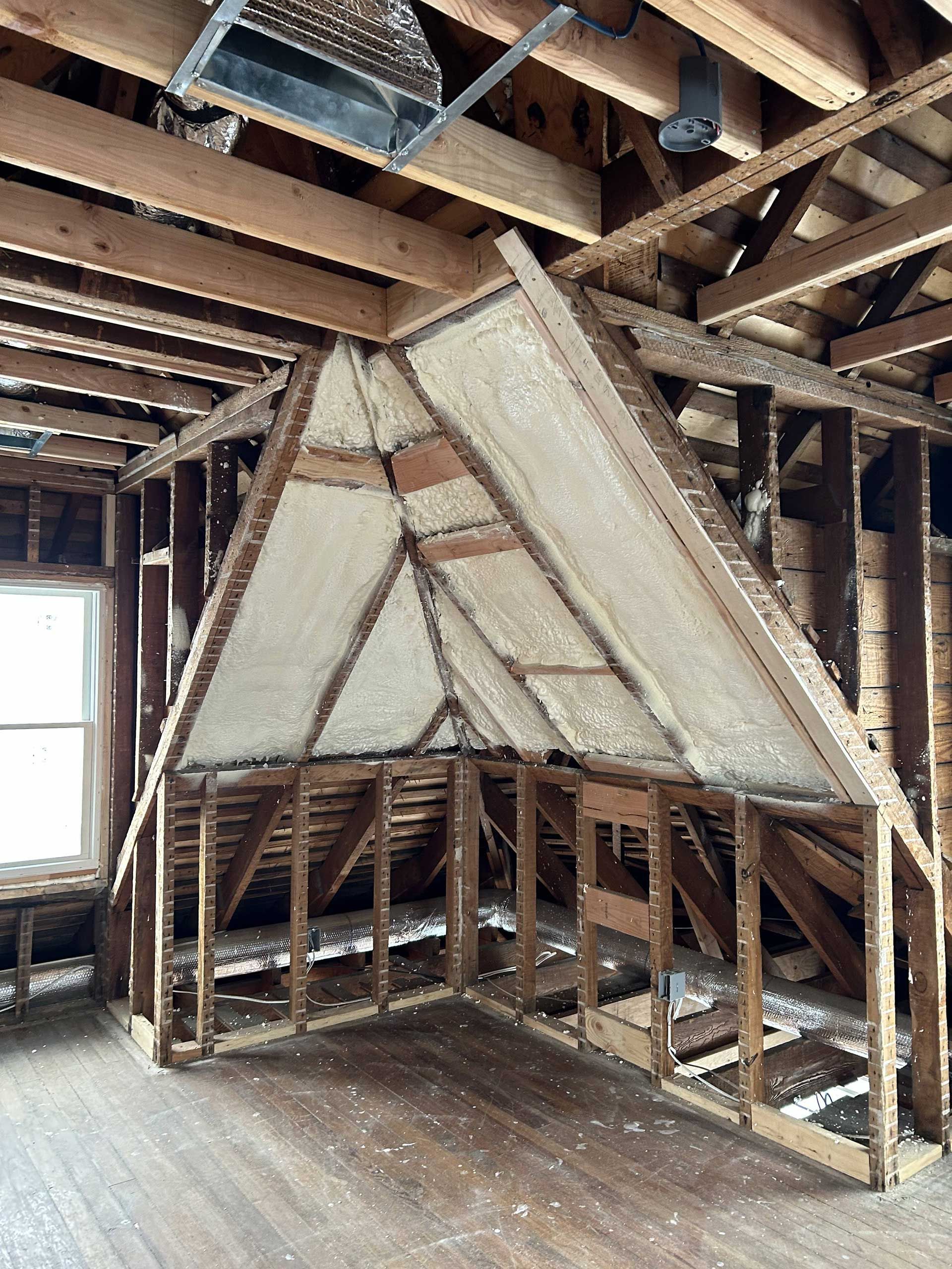 An unfinished attic space showing exposed wood framing and triangular wall sections covered in off-white insulation.