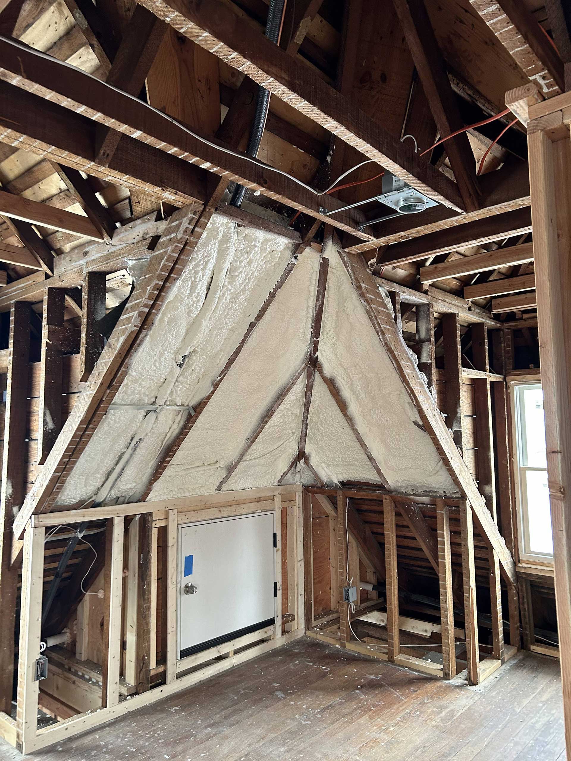 An unfinished attic renovation with exposed wooden framing and triangular panels insulated with white spray foam.
