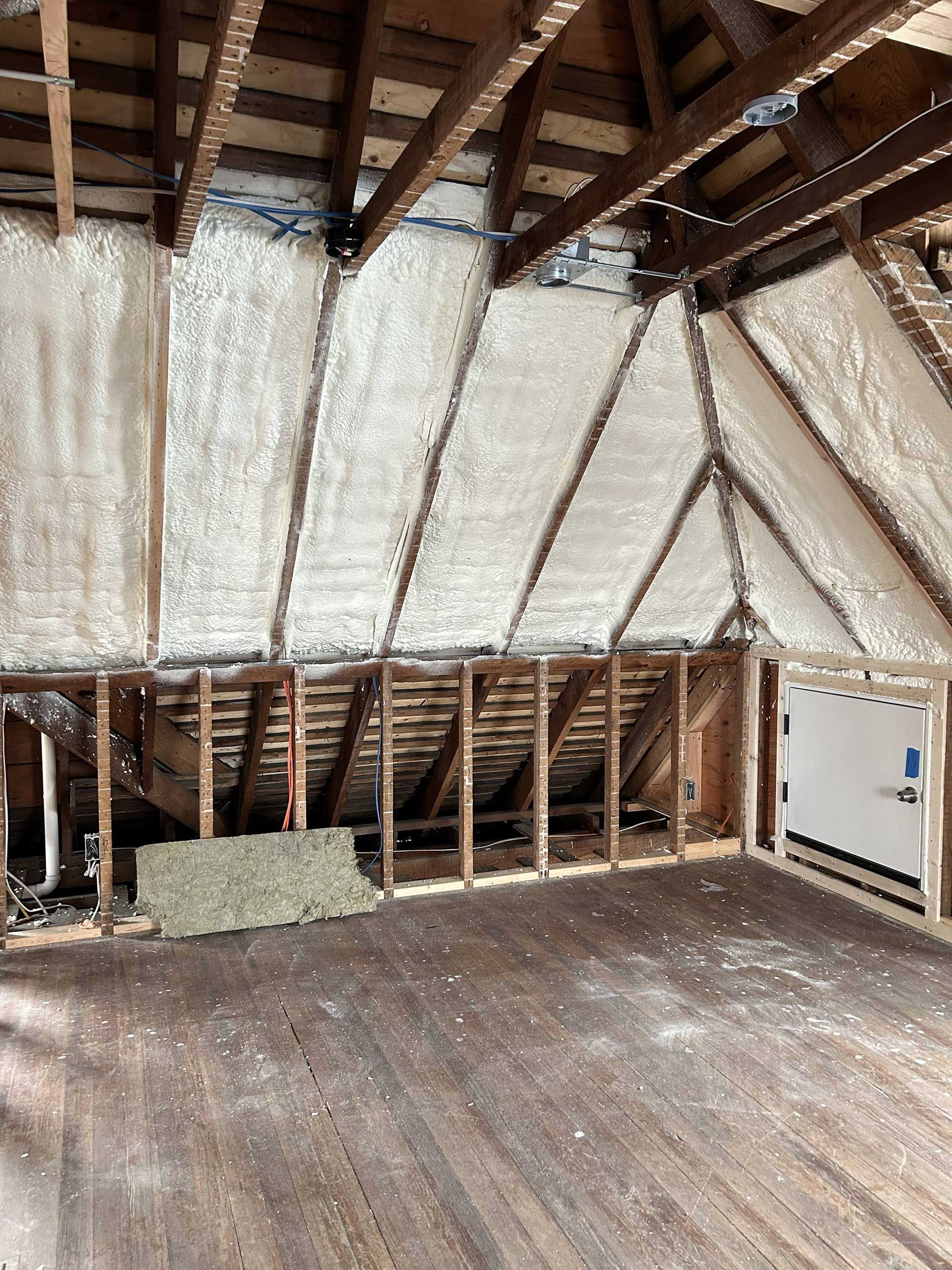 An unfinished attic with exposed wooden rafters, white spray foam insulation between beams, and wood flooring.