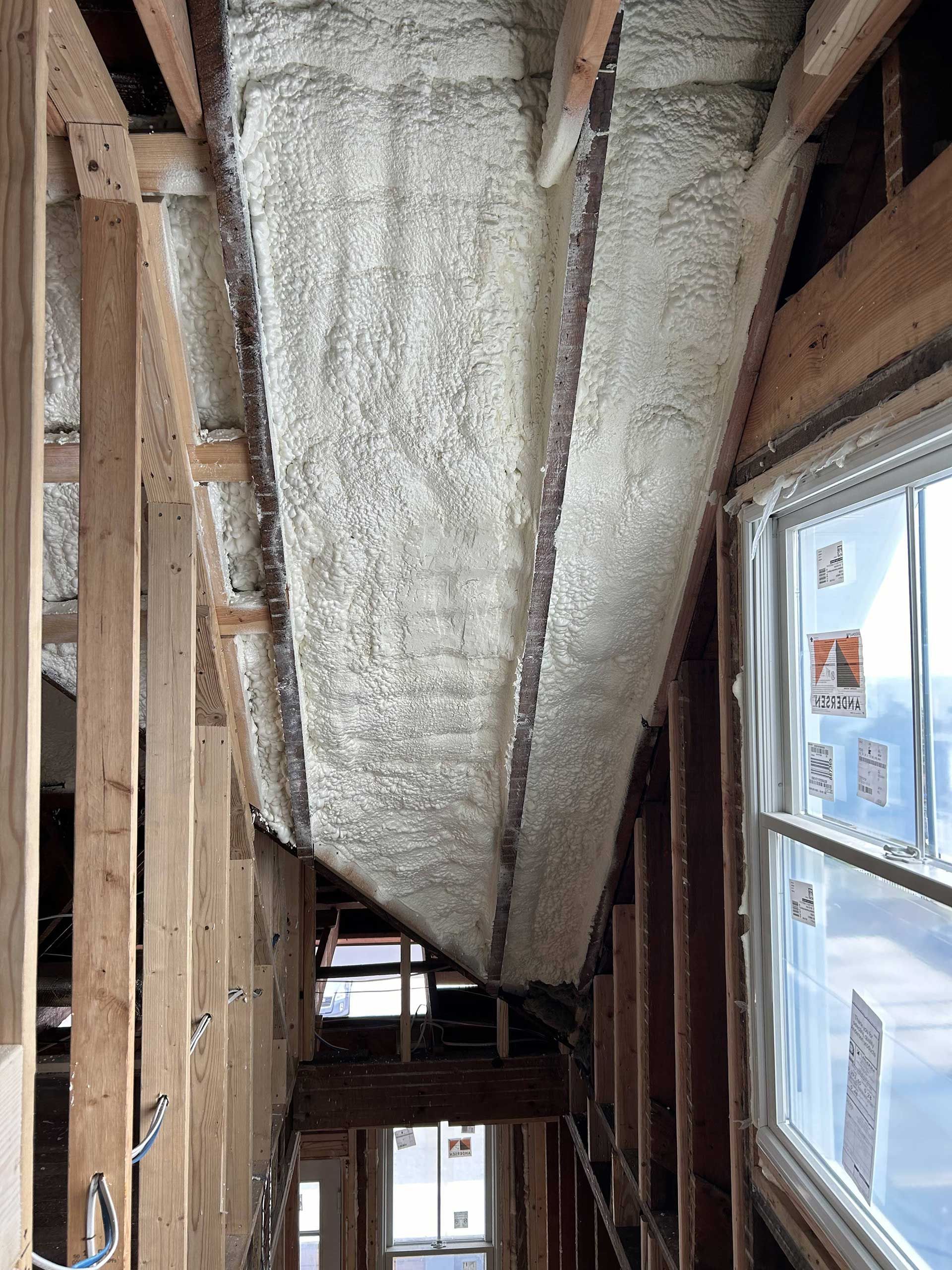 A view of a building under construction featuring spray foam insulation applied between exposed wooden wall studs.