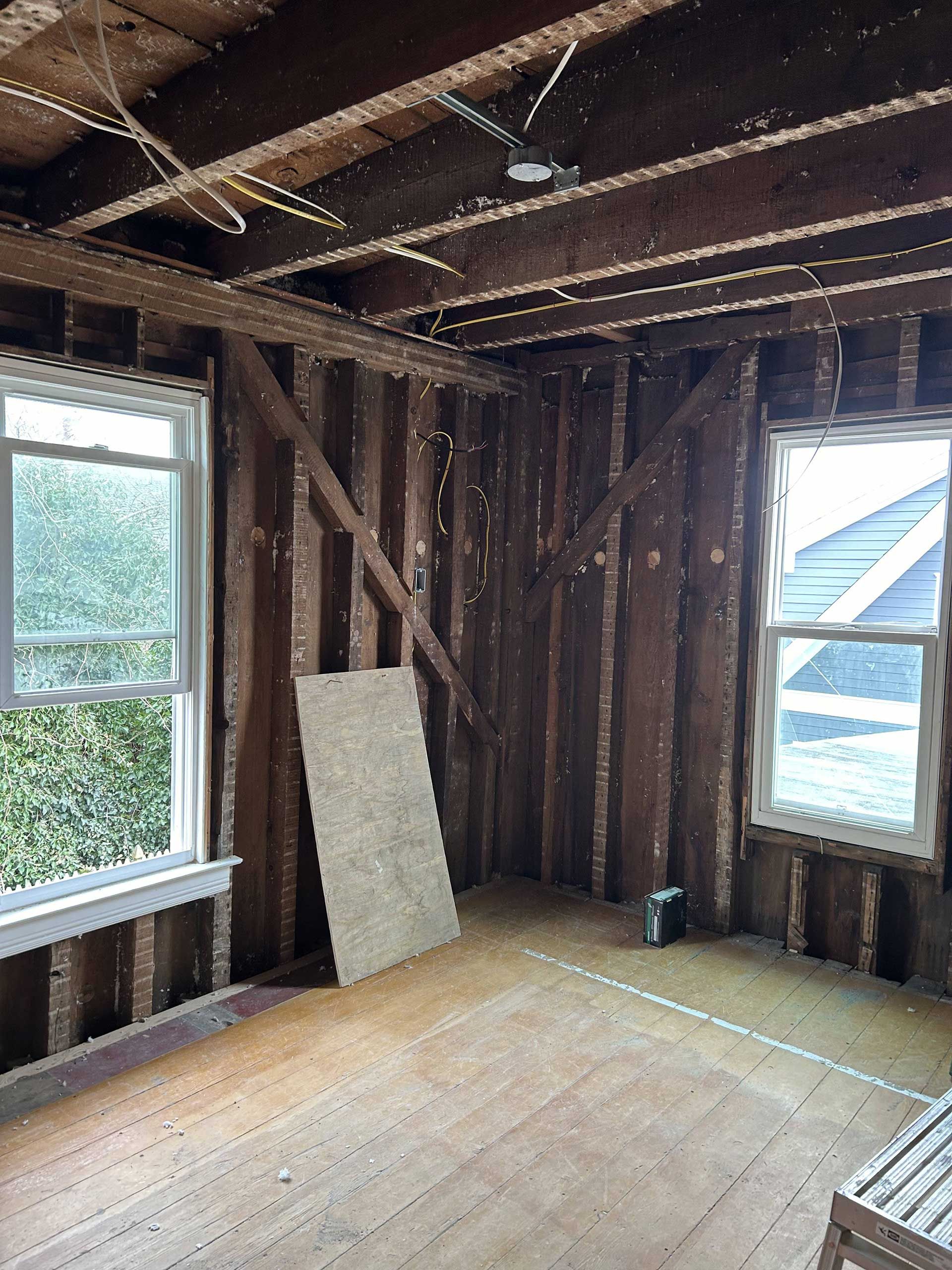 An interior view of a room under construction, featuring exposed wooden wall studs, floorboards, and two windows.