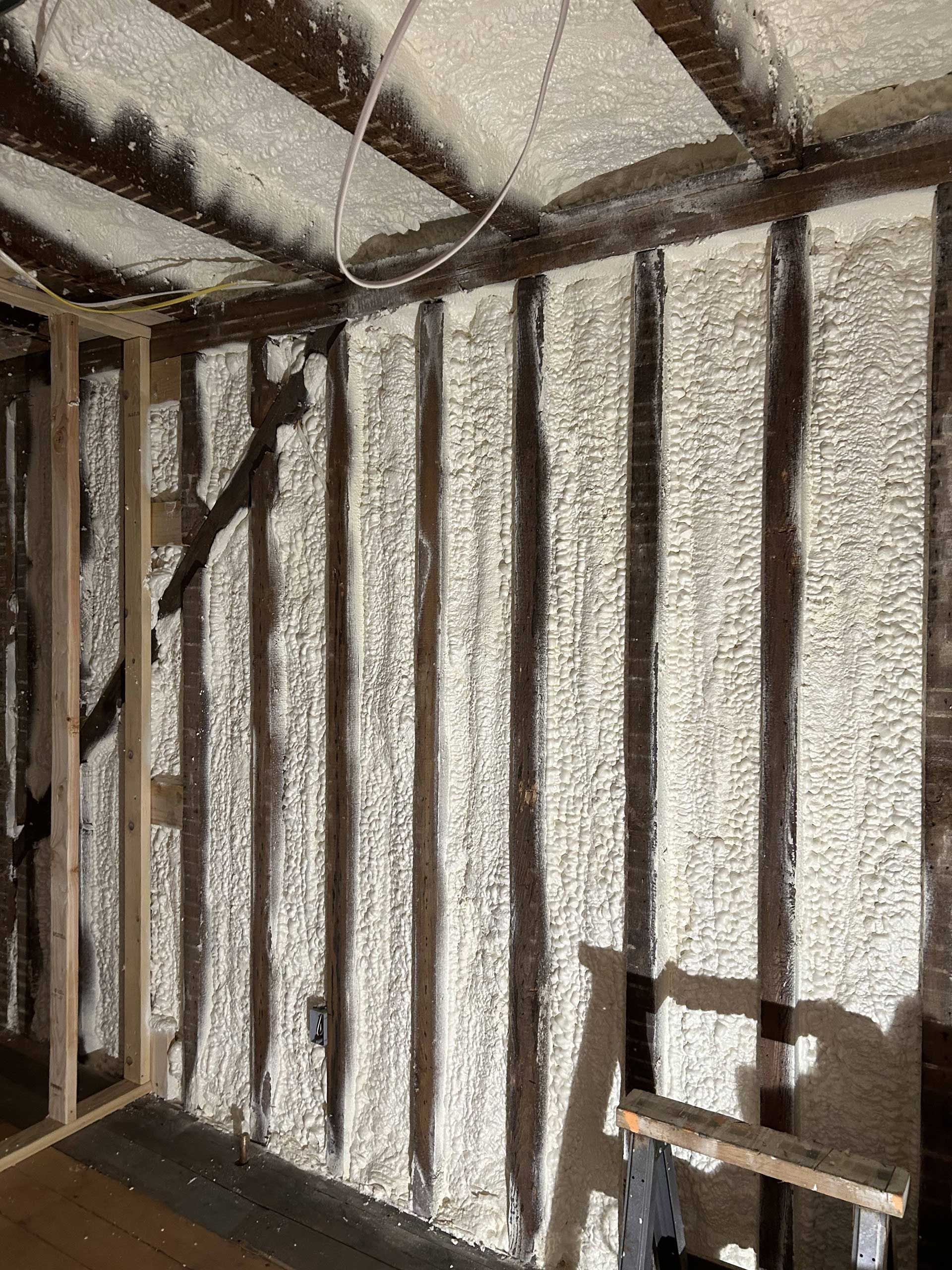 The interior of a building under renovation, showing dark wooden wall studs insulated with white spray foam.