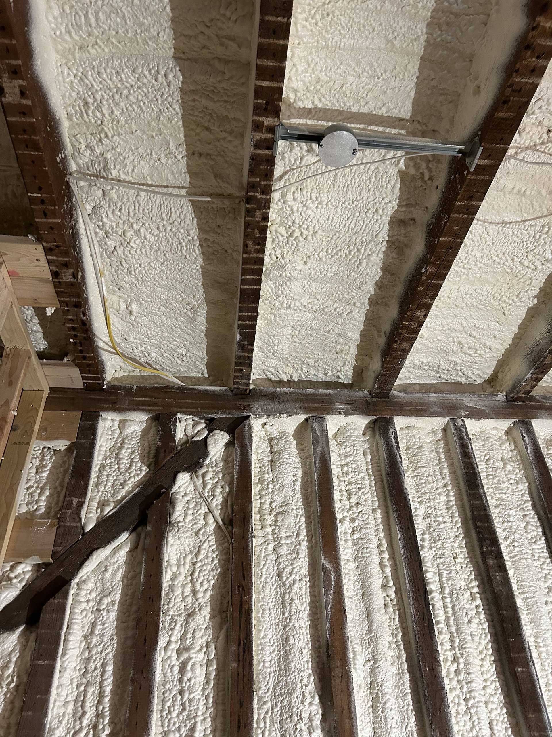 Exposed dark wooden ceiling beams between sections of white spray foam insulation in a construction site.