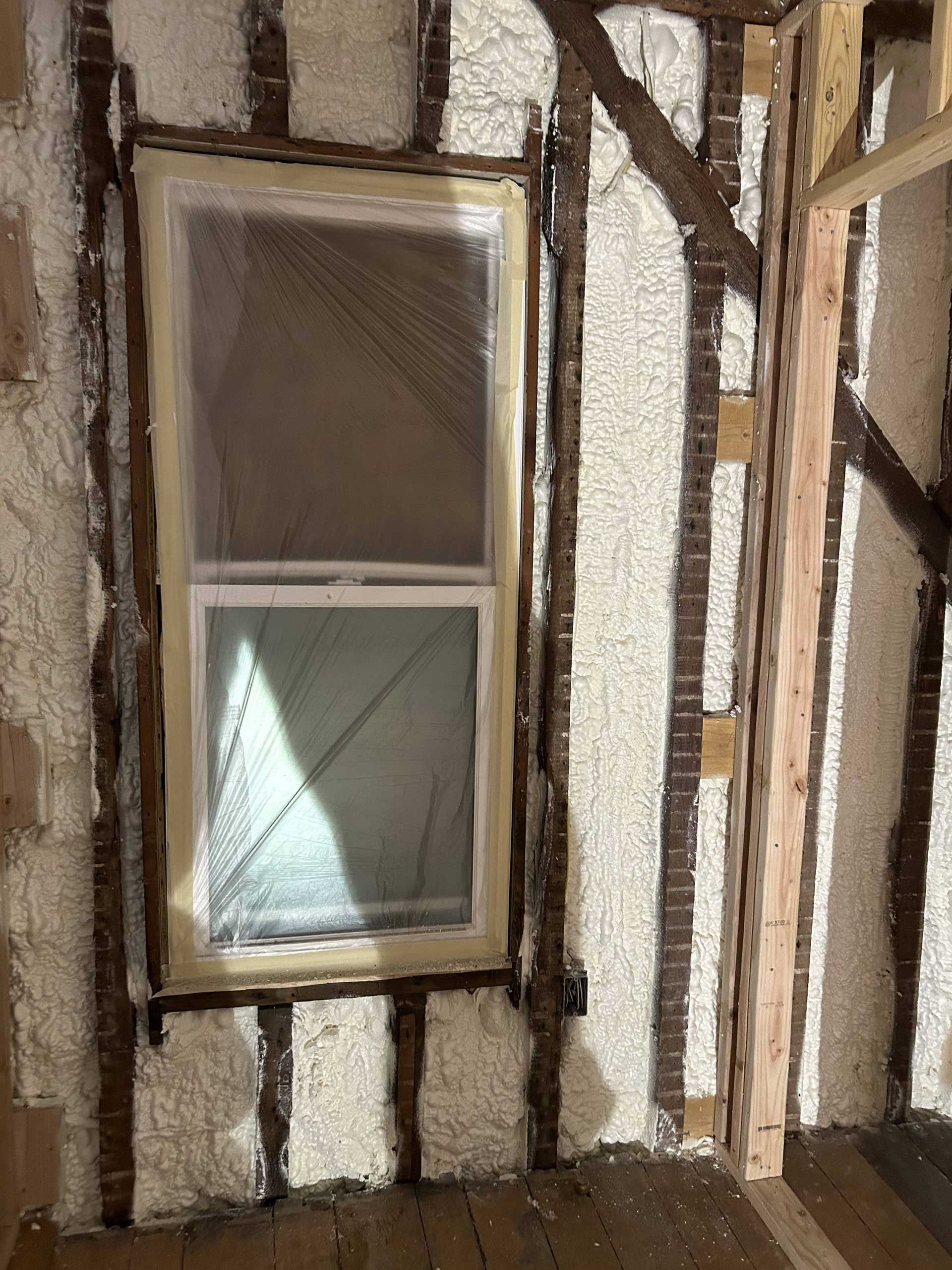 Interior construction scene showing a window frame installed between timber wall studs, filled with white spray foam insulation.