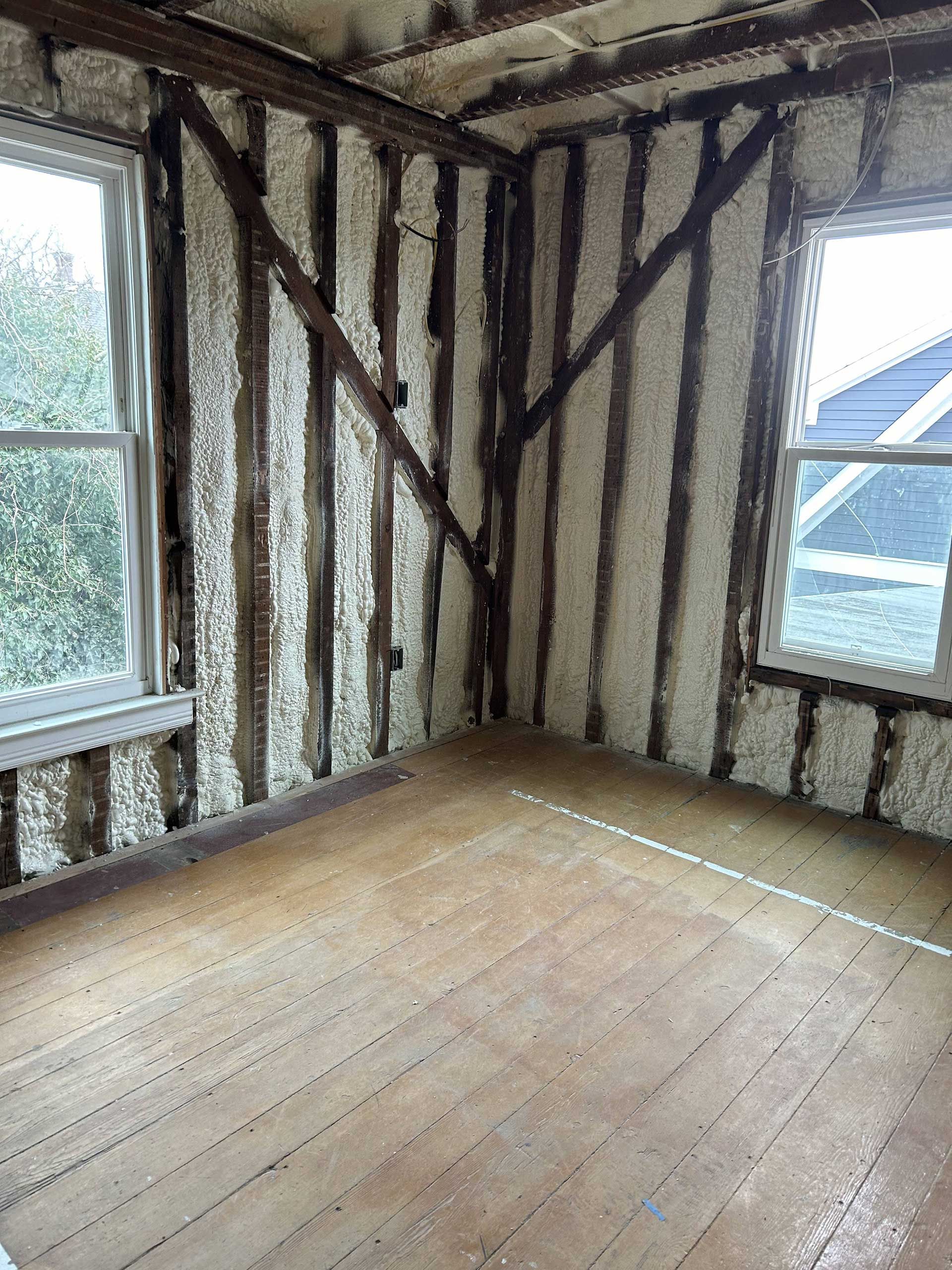An unfinished room with exposed wooden wall framing and floorboards, filled with white spray foam insulation.