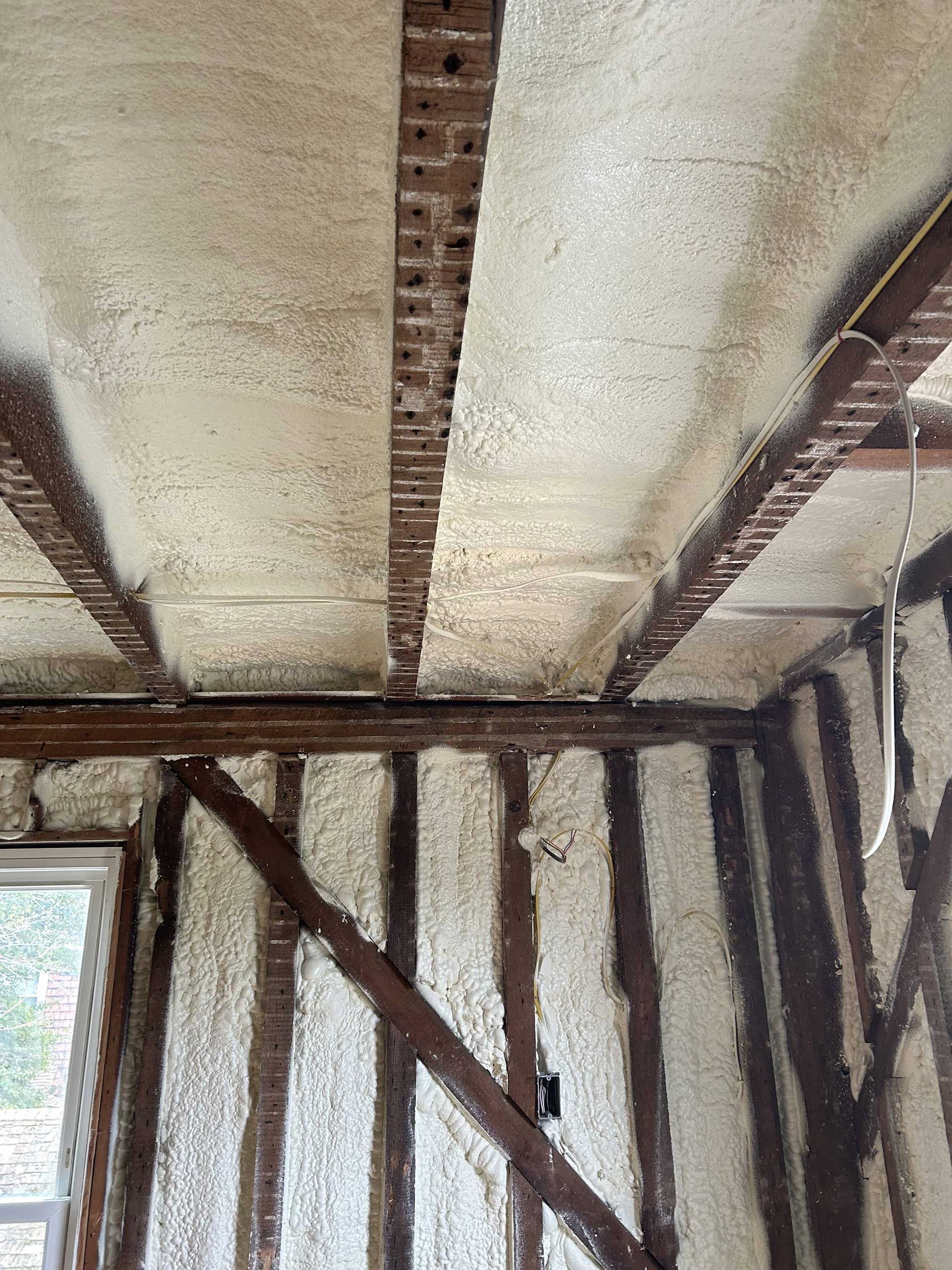 Walls and ceiling of a timber-framed room insulated with white spray foam between exposed dark wooden beams and studs.