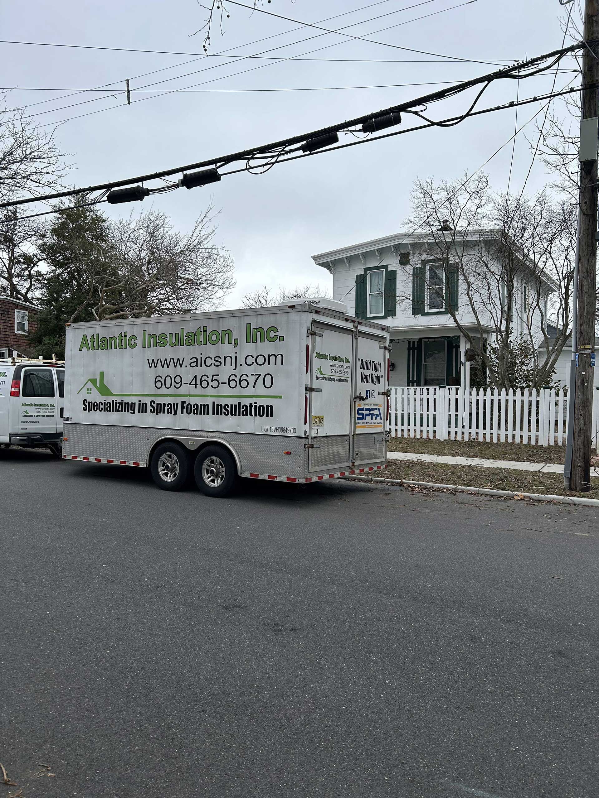 An Atlantic Insulation, Inc. work trailer parked on a residential street next to a white house with a picket fence.