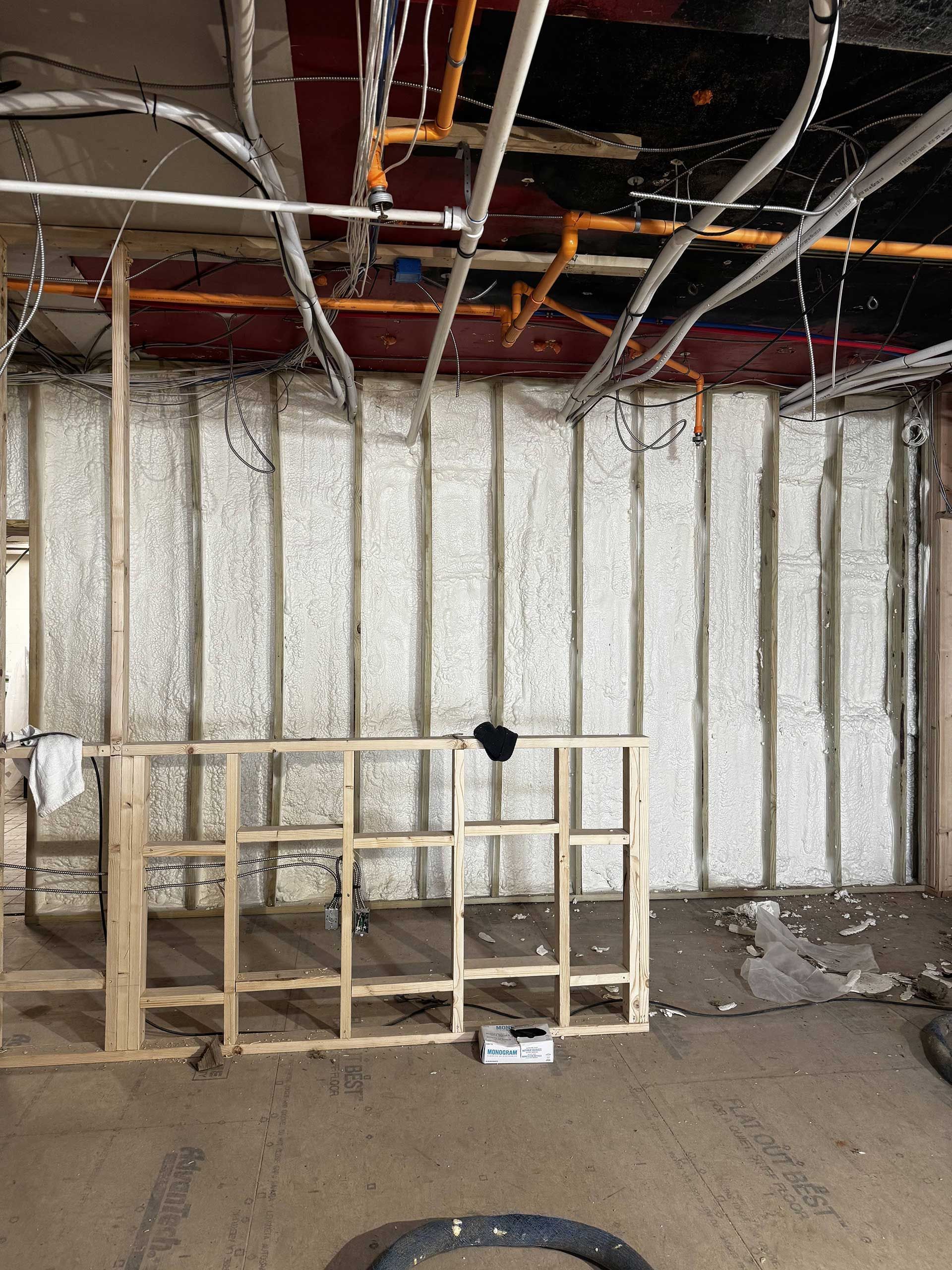 A construction scene showing an unfinished room with exposed wall studs, white spray foam insulation, and open ceiling pipes.
