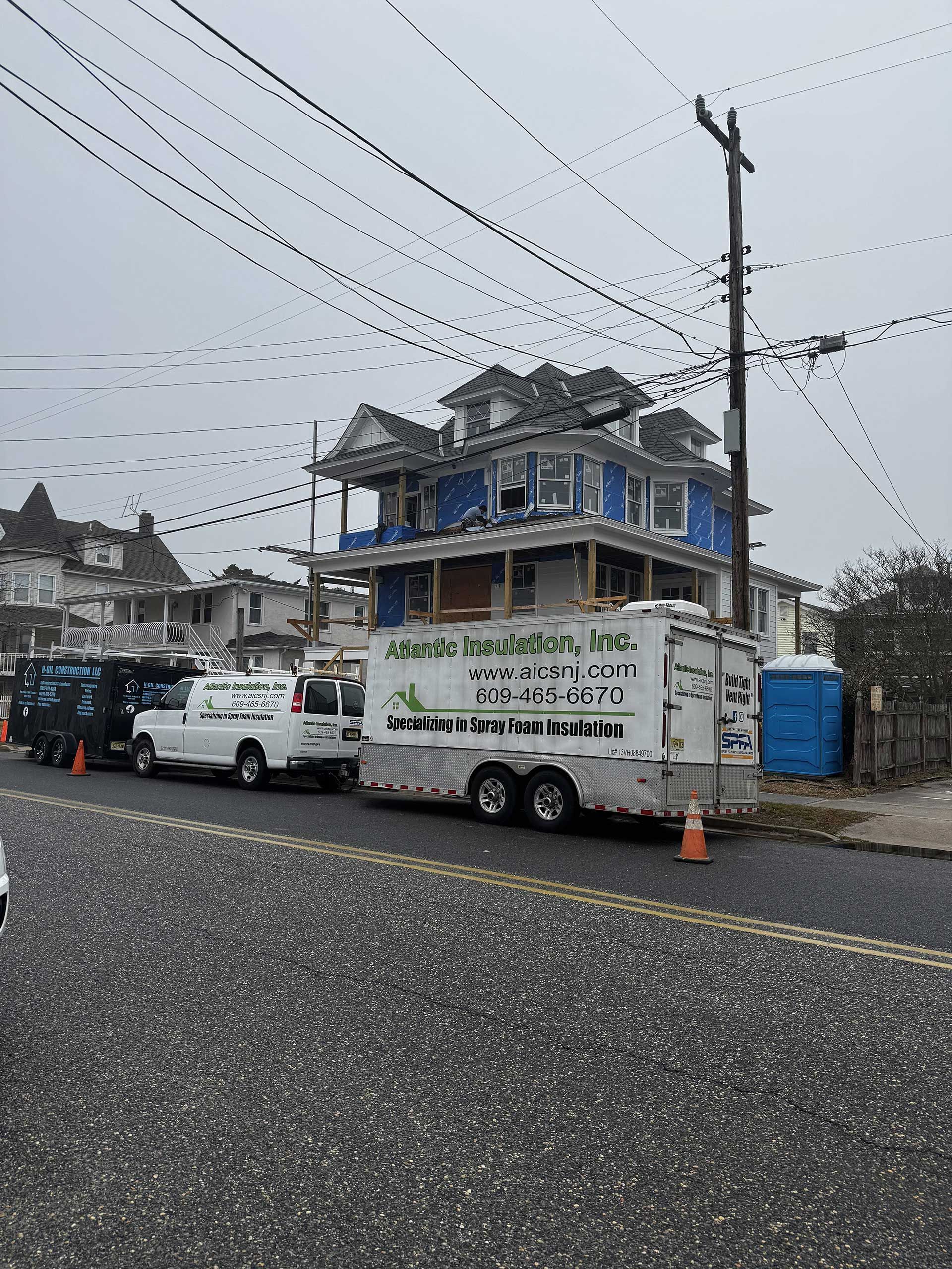A white trailer and van parked on a street in front of a blue, multi-story house undergoing renovations.