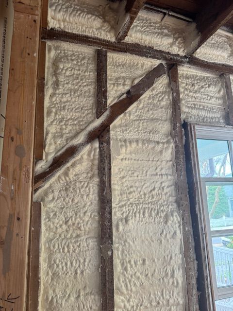 Interior wall insulated with light beige spray foam, covering wood studs. Window visible on right.