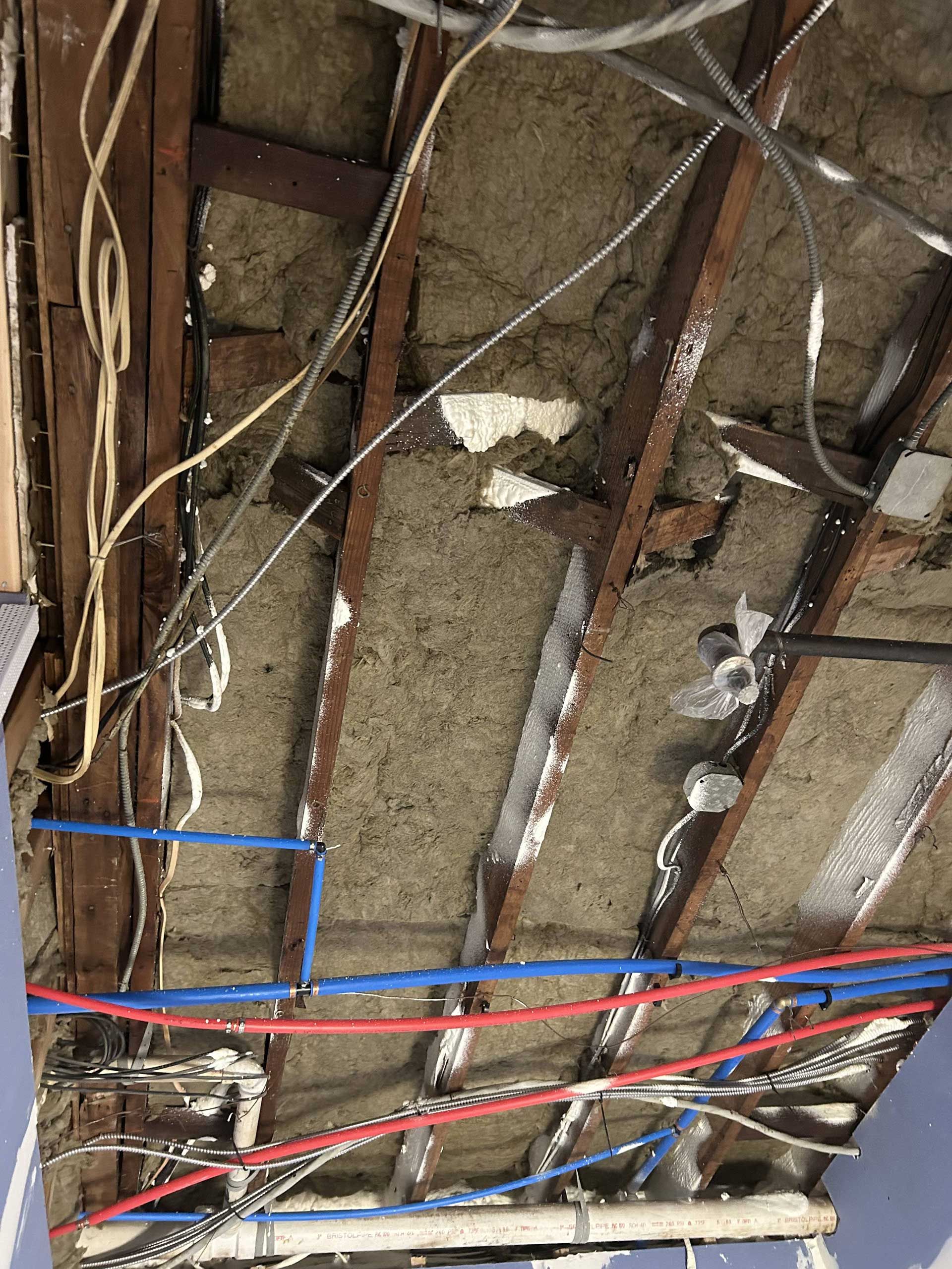 Exposed ceiling framing with red and blue PEX plumbing pipes, electrical wiring, and insulation between wooden joists.
