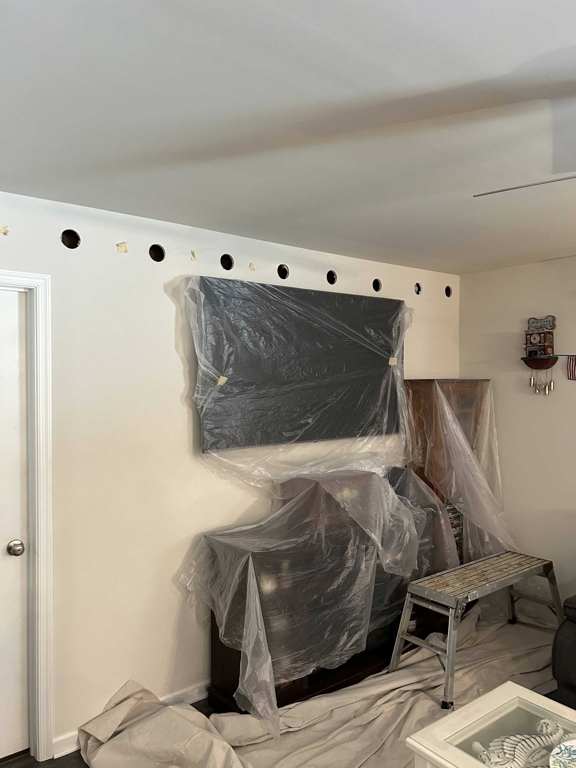 A wall in a room undergoing construction, featuring a line of circular holes near the ceiling and furniture covered in plastic.