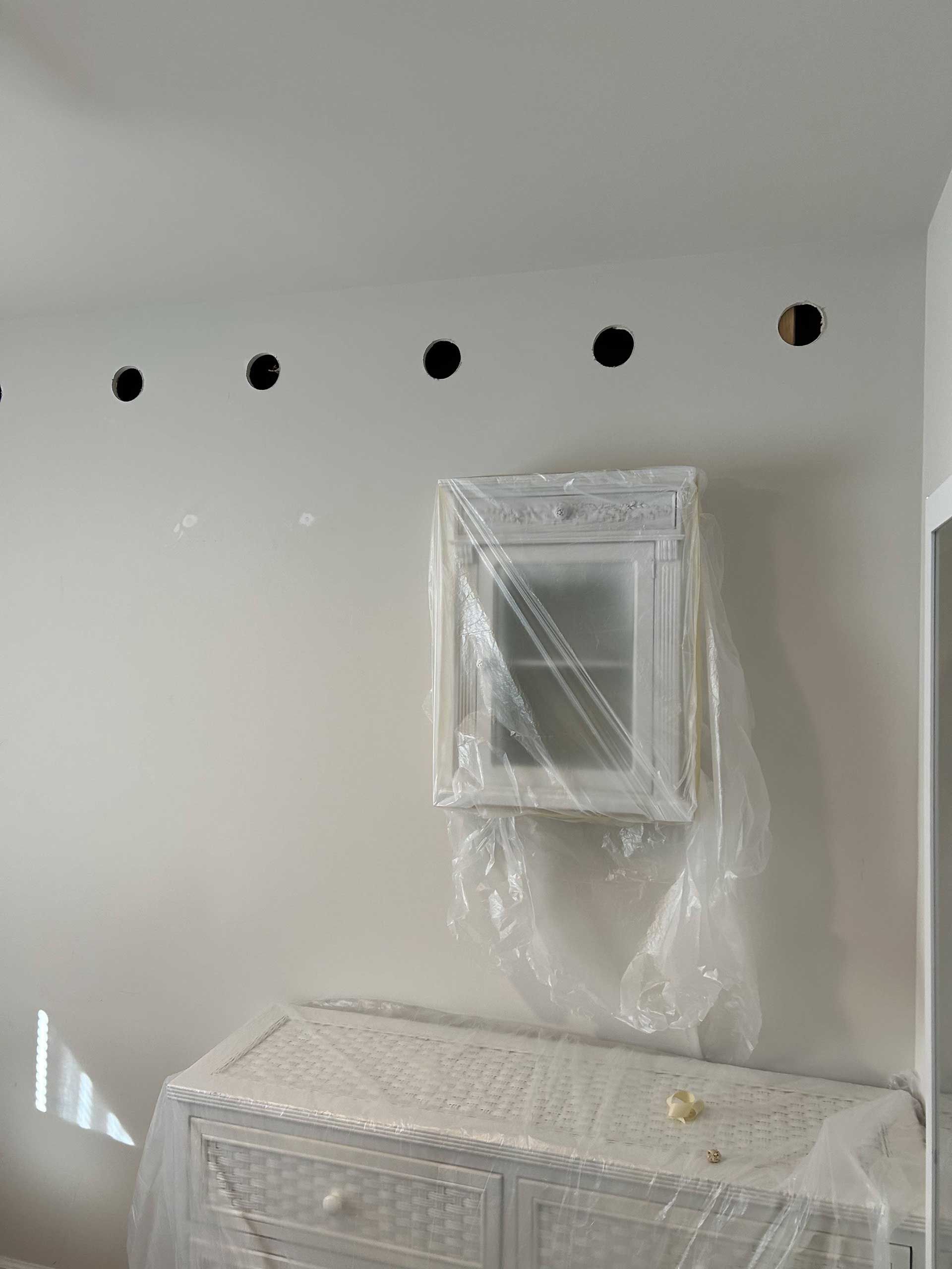 A room under renovation with five evenly spaced holes in the wall above a cabinet and dresser covered in plastic sheets.