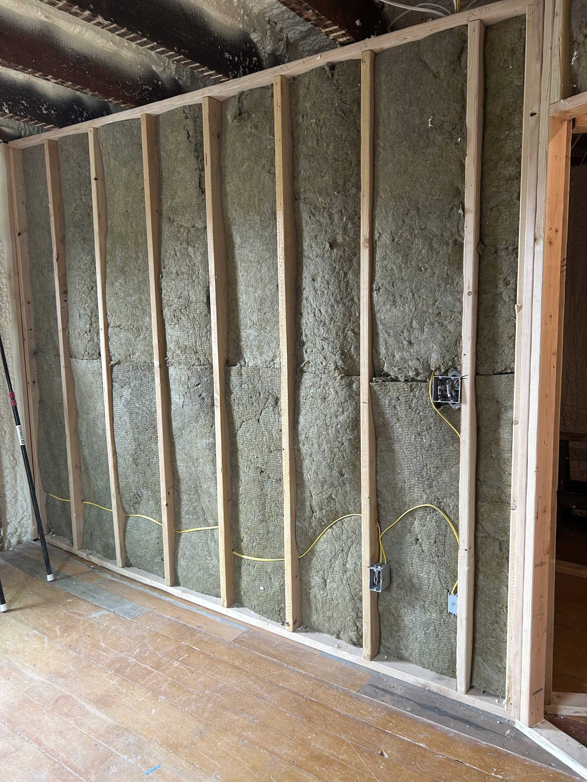 A wall frame interior construction showing wood studs, grey insulation batts, and electrical wiring.