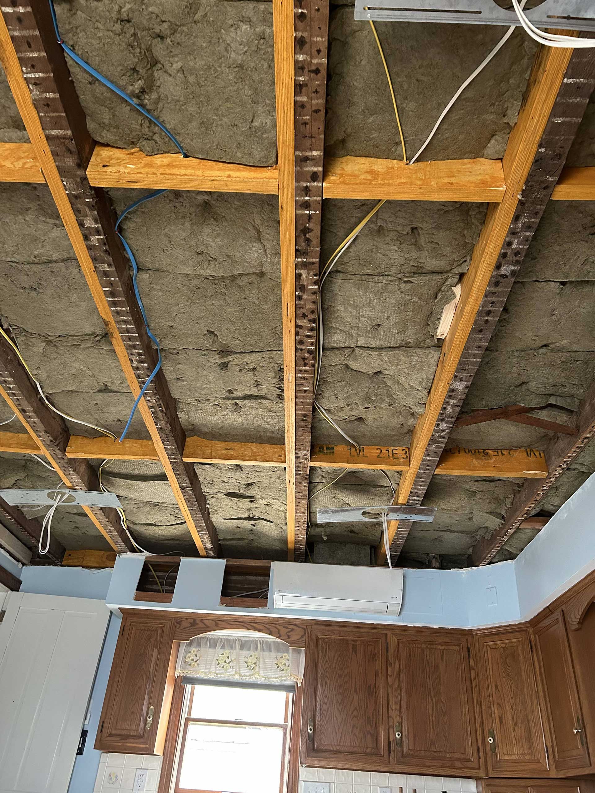 An unfinished ceiling with exposed wooden joists, fibrous insulation, and electrical wiring above a kitchen cabinet wall.