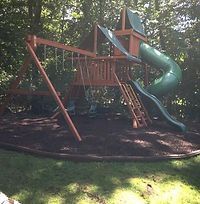 A wooden swing set with a green slide and swings in a backyard.