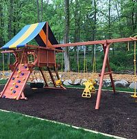 A wooden playground set with a climbing wall and swings in a backyard.