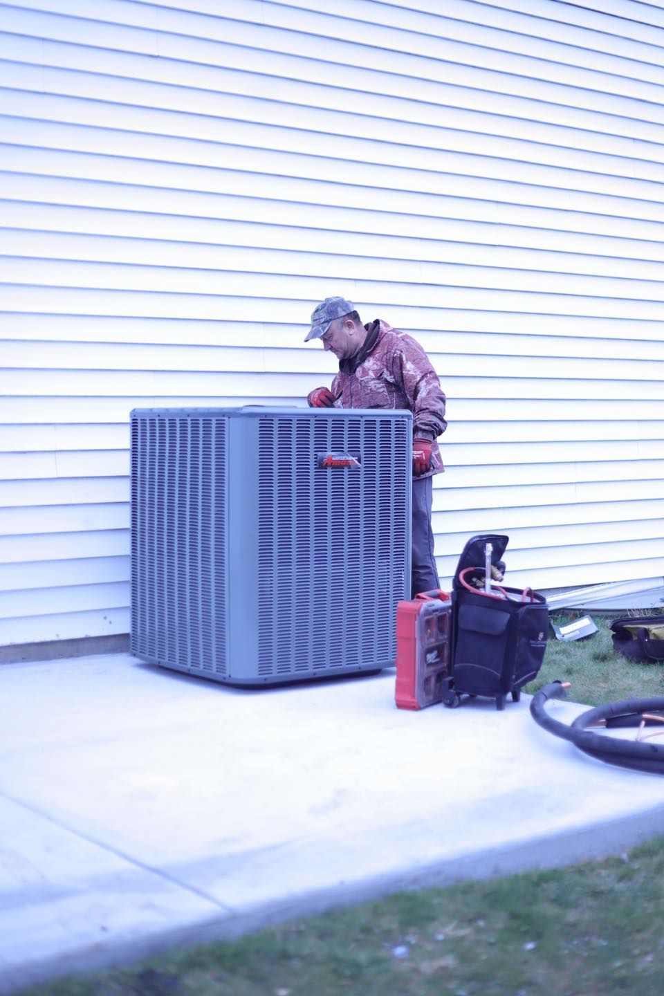 Person working on an outdoor HVAC unit beside a house, with tools and hose on the ground.