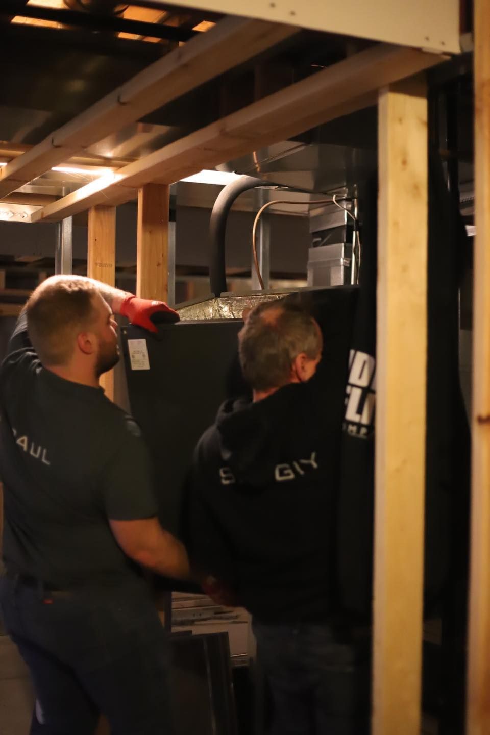Two workers in black shirts moving a large appliance in a wooden-framed basement space