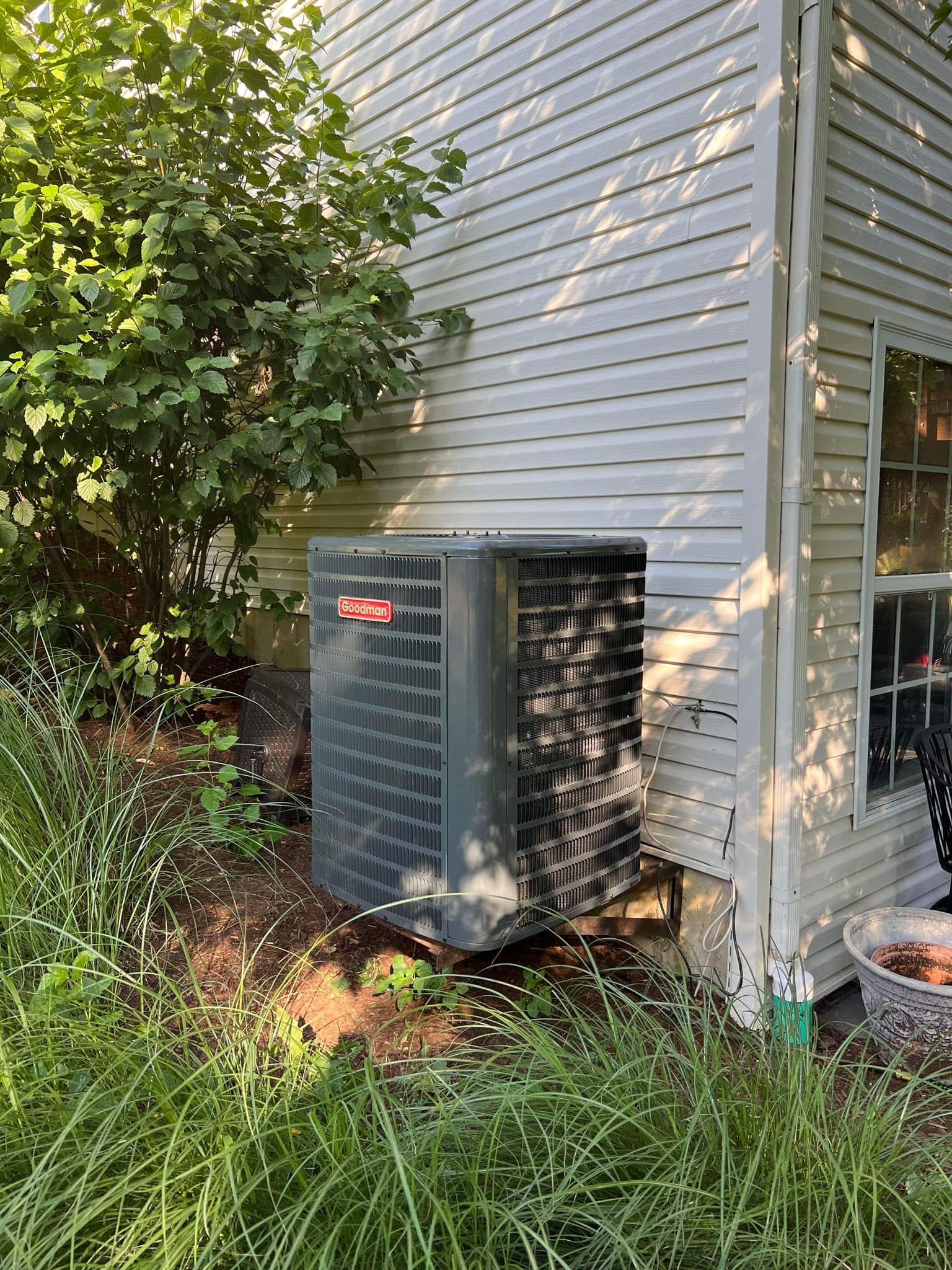 Outdoor air conditioner unit beside a house amid tall grass and shrubs