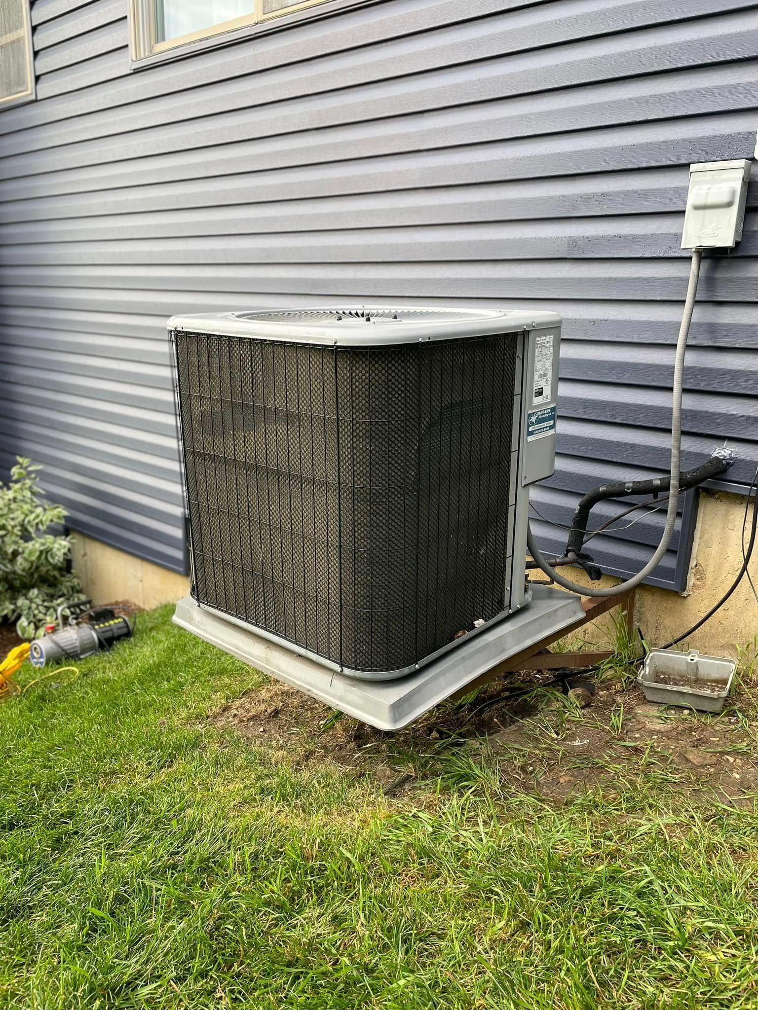 Outdoor HVAC condenser unit beside a gray house wall on a metal pad, with grass and utility wiring nearby