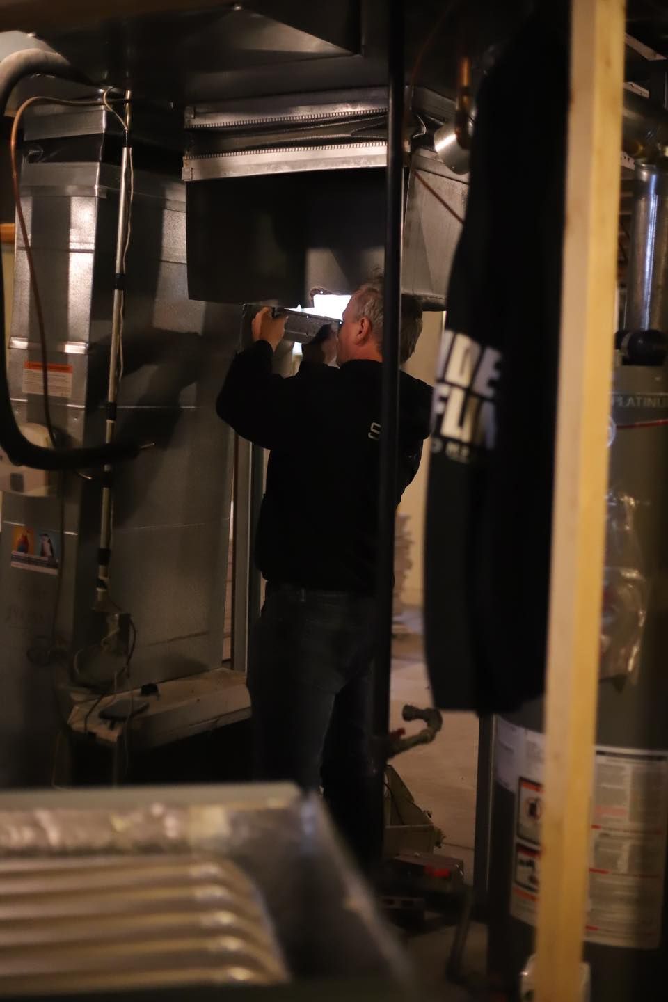 Person inspecting ductwork in a dimly lit utility room, wearing a black shirt and jeans.
