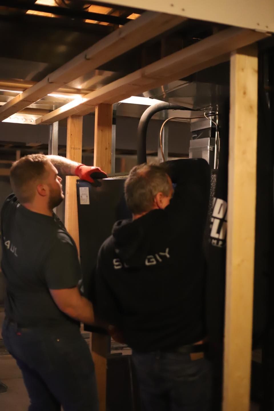 Two workers installing a machine in a wooden-framed basement, one in red gloves pointing upward.