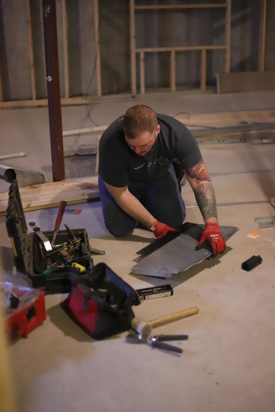 Person kneels on a concrete floor, cutting sheet metal with a saw amid tools in a workshop under construction