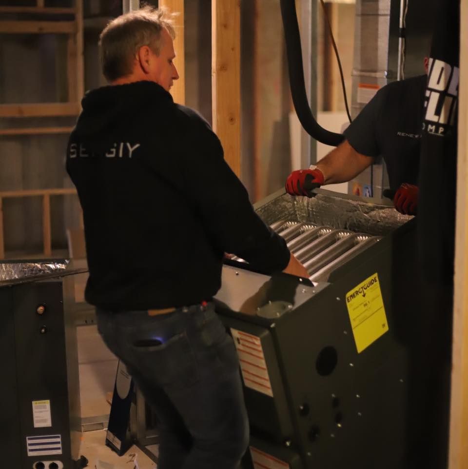 Two workers installing a large metal HVAC unit in an unfinished room.