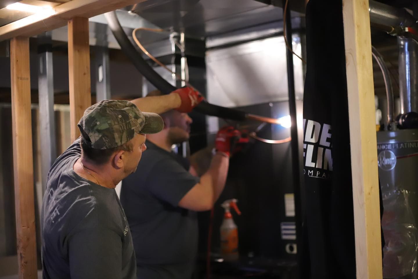 Two workers in gloves install ductwork in a basement with exposed framing and insulation.