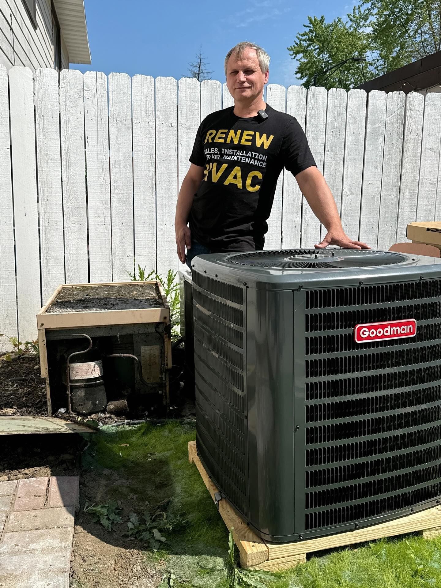 Man standing beside a large outdoor air conditioner unit in a fenced backyard, wearing a black “RENEW HVAC” shirt.