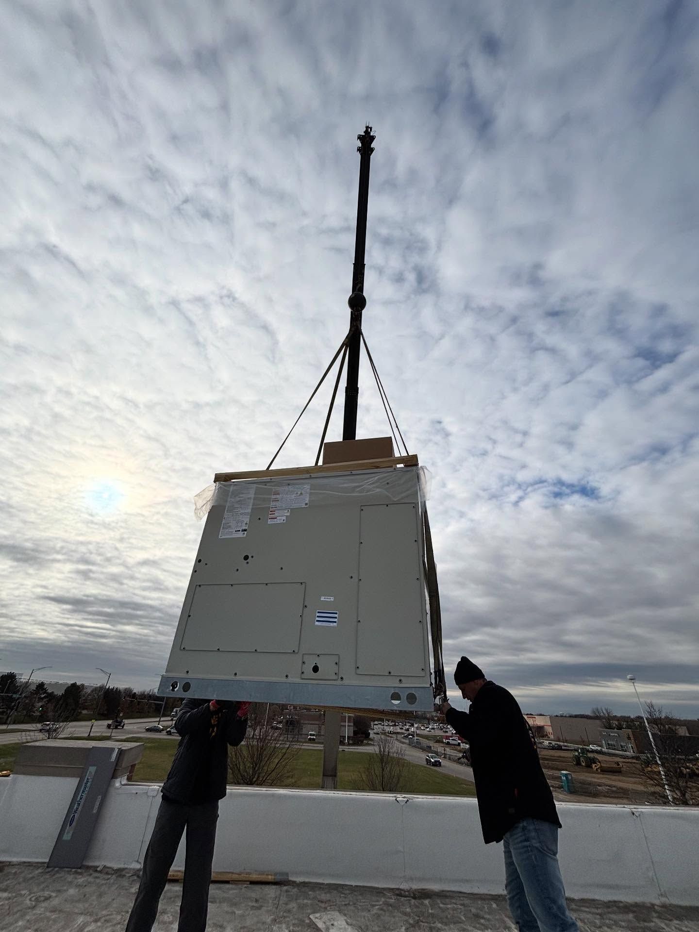 Two workers hoist a large metal unit with a crane on a rooftop under a cloudy sky.