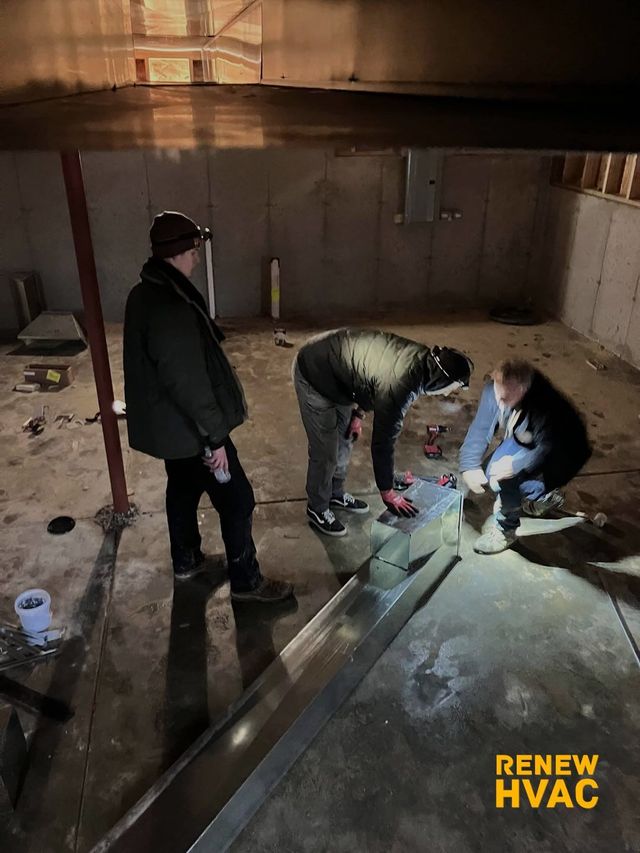 Three people inspecting and repairing a floor in a dim wooden basement with tools and a flashlight.