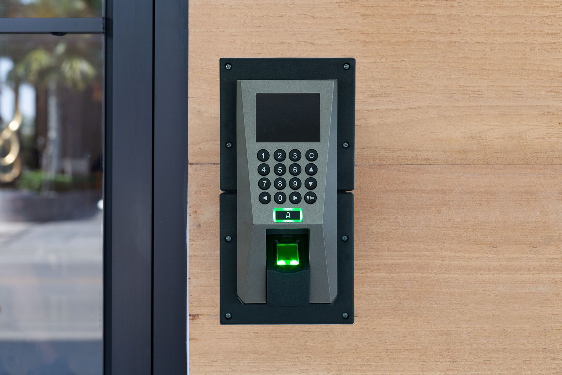 Wall-mounted keypad access control panel with card reader on a wood wall beside a glass door.