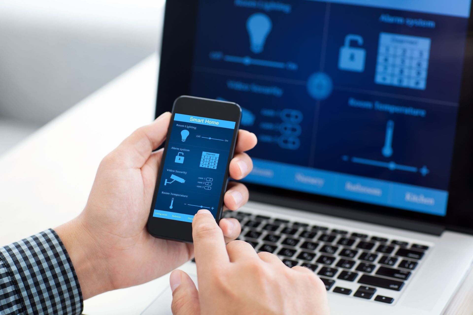 Hands holding a smartphone with smart home controls in front of a laptop dashboard