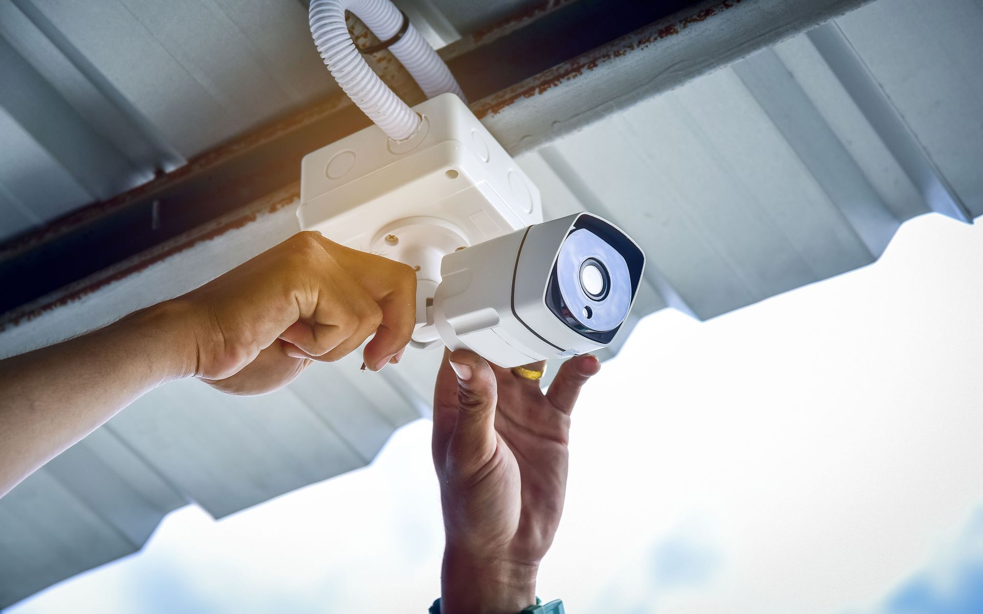 Hands adjusting a white security camera mounted under a corrugated metal roof.