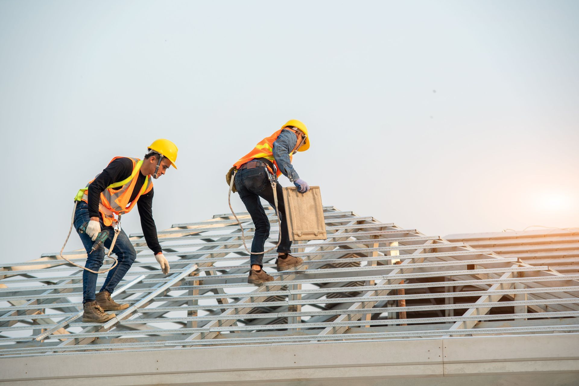 Two construction workers in safety gear and hard hats installing metal roof framing against a bright sky.