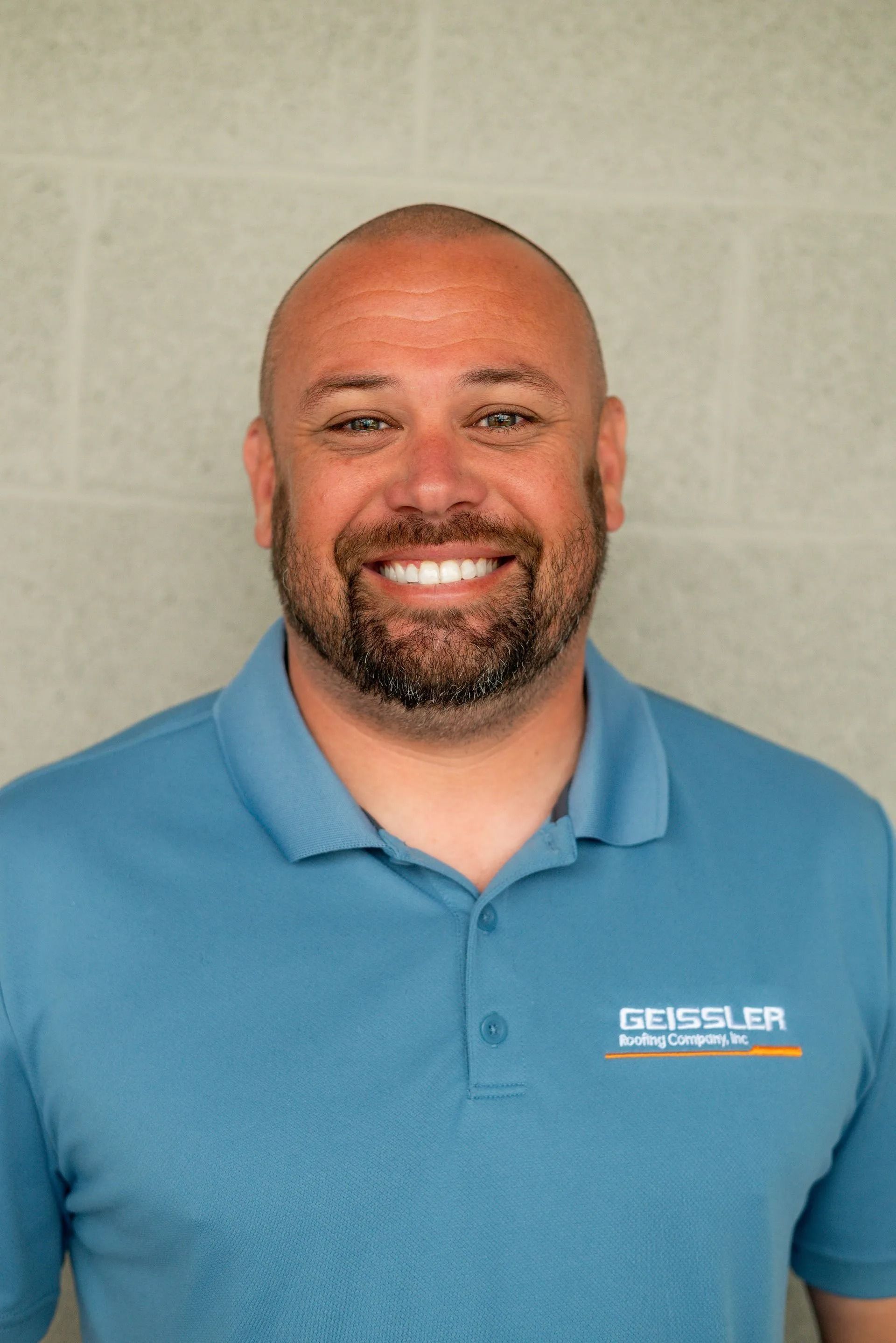 A smiling man in a blue polo shirt featuring the Gessler logo against a grey brick wall.