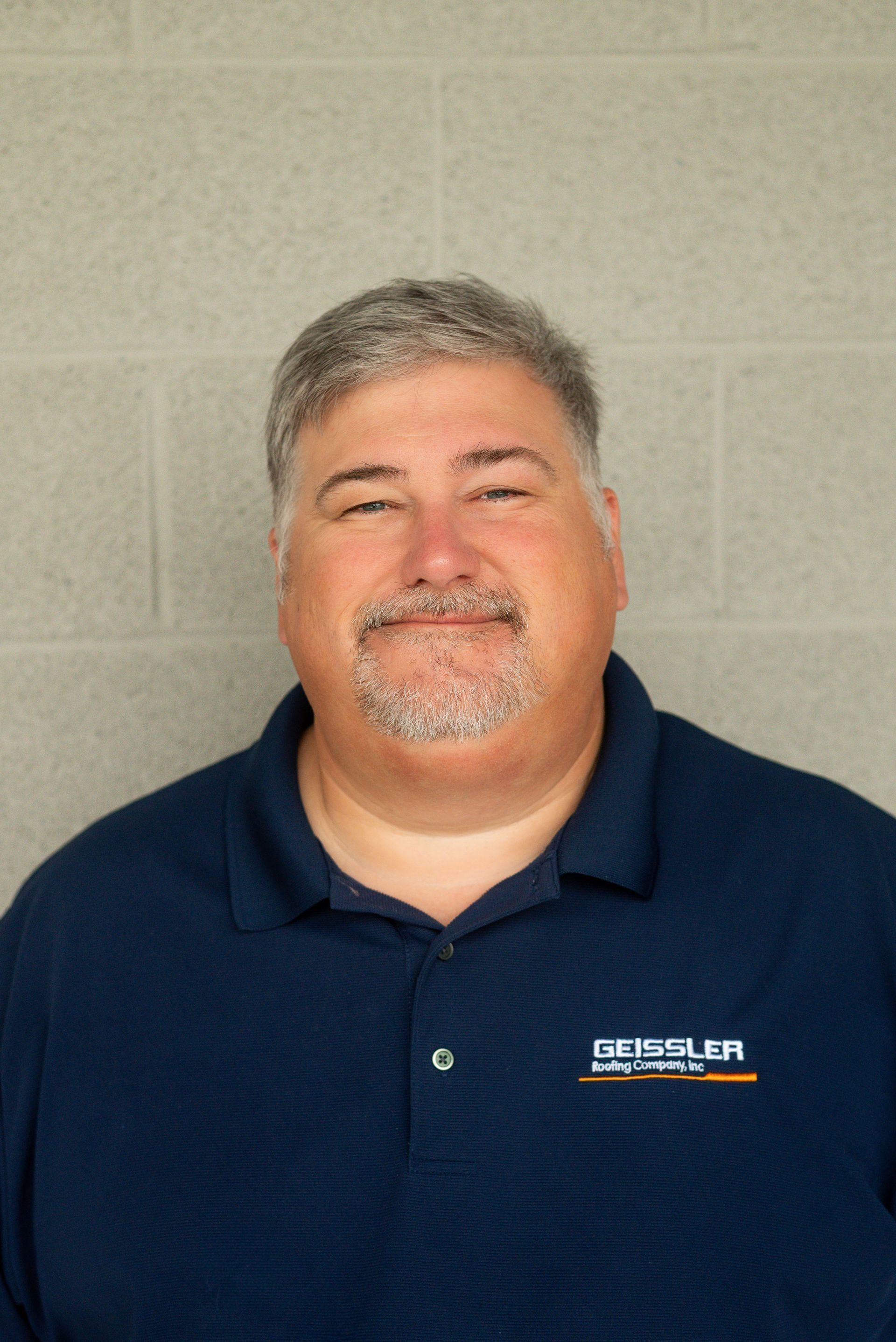 A man with grey hair and a goatee smiles against a grey brick wall, wearing a navy blue polo shirt with a company logo.